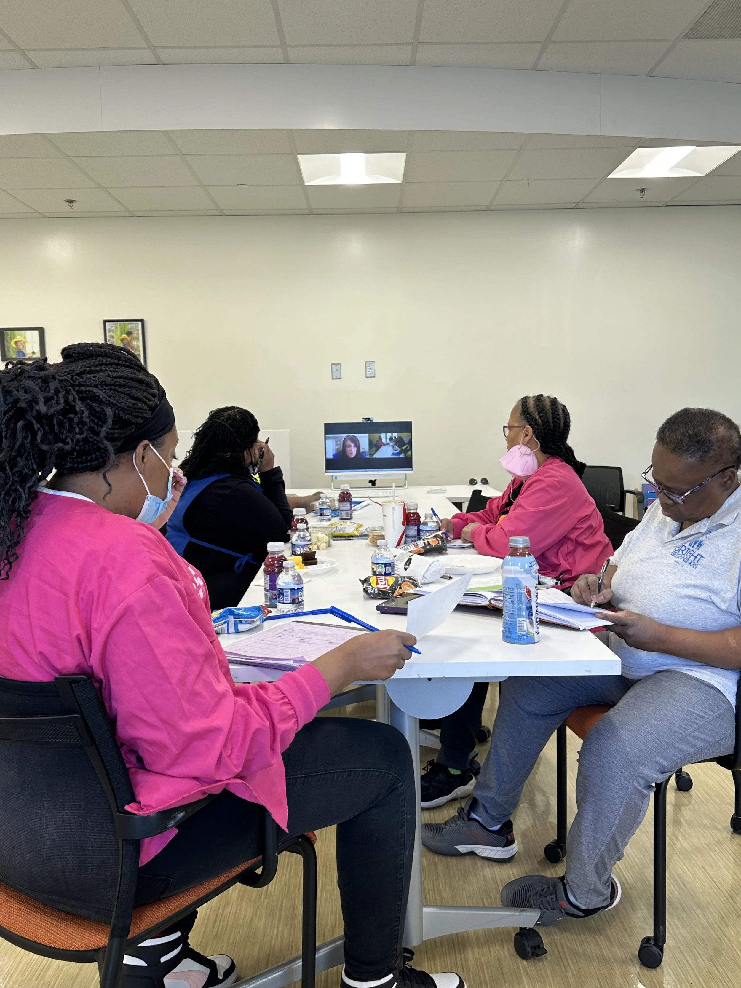 A group of five people, some wearing pink masks and scrubs, are sitting around a table in a conference room, attending a virtual meeting on a computer.