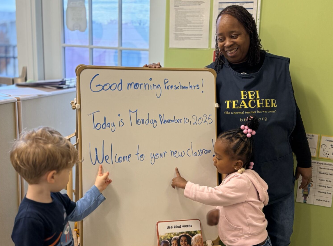 A teacher and two young children standing near a whiteboard in a classroom. The whiteboard has a message welcoming students on their first day of school, Monday, November 16, 2025. The teacher is smiling and wearing a black vest with yellow text. The children are pointing at the whiteboard, one with a blue shirt and the other in a pink hoodie.