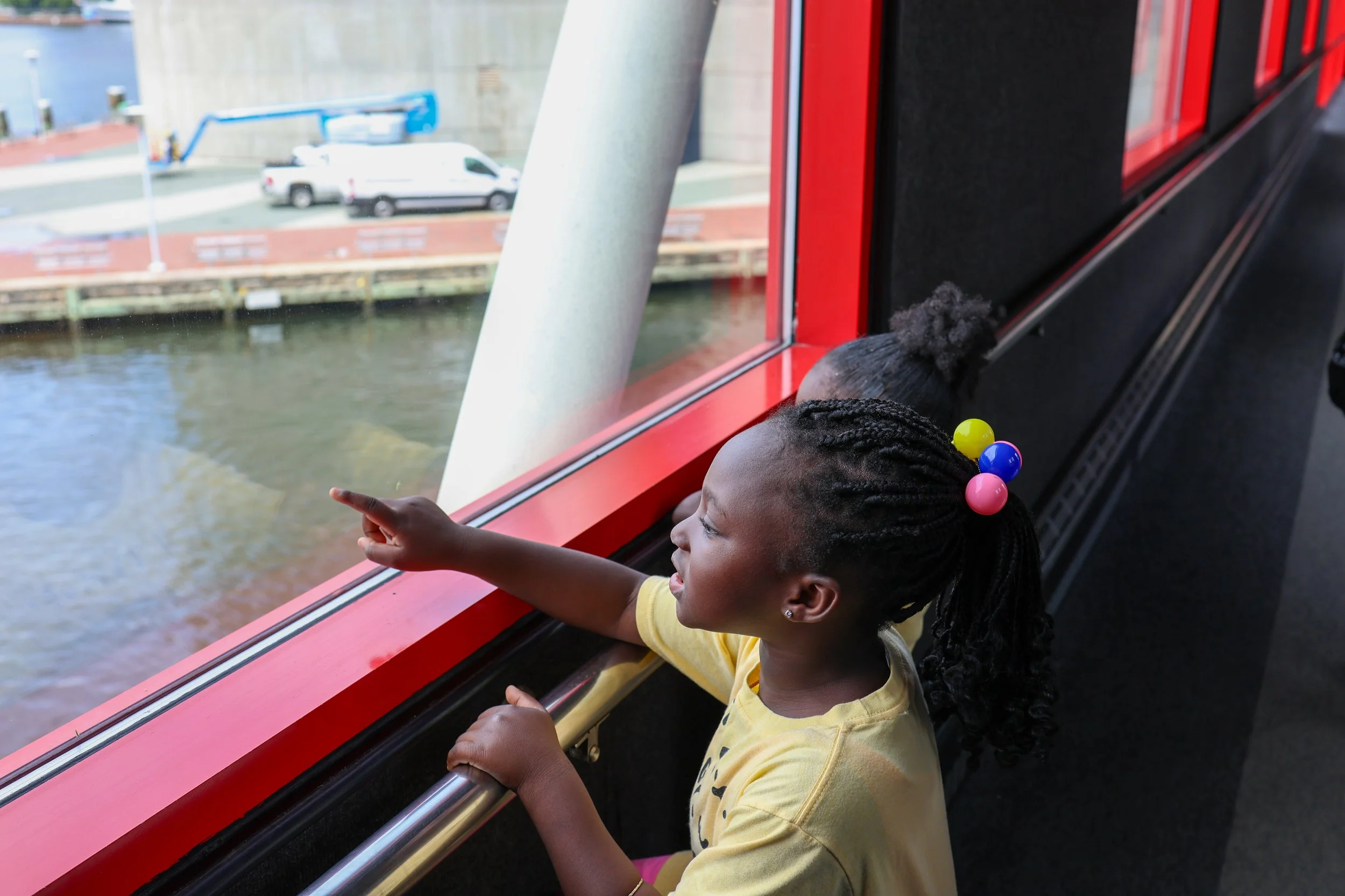 A young girl with braided hair pointing out of a window on a boat or train, with a view of the river and buildings outside.