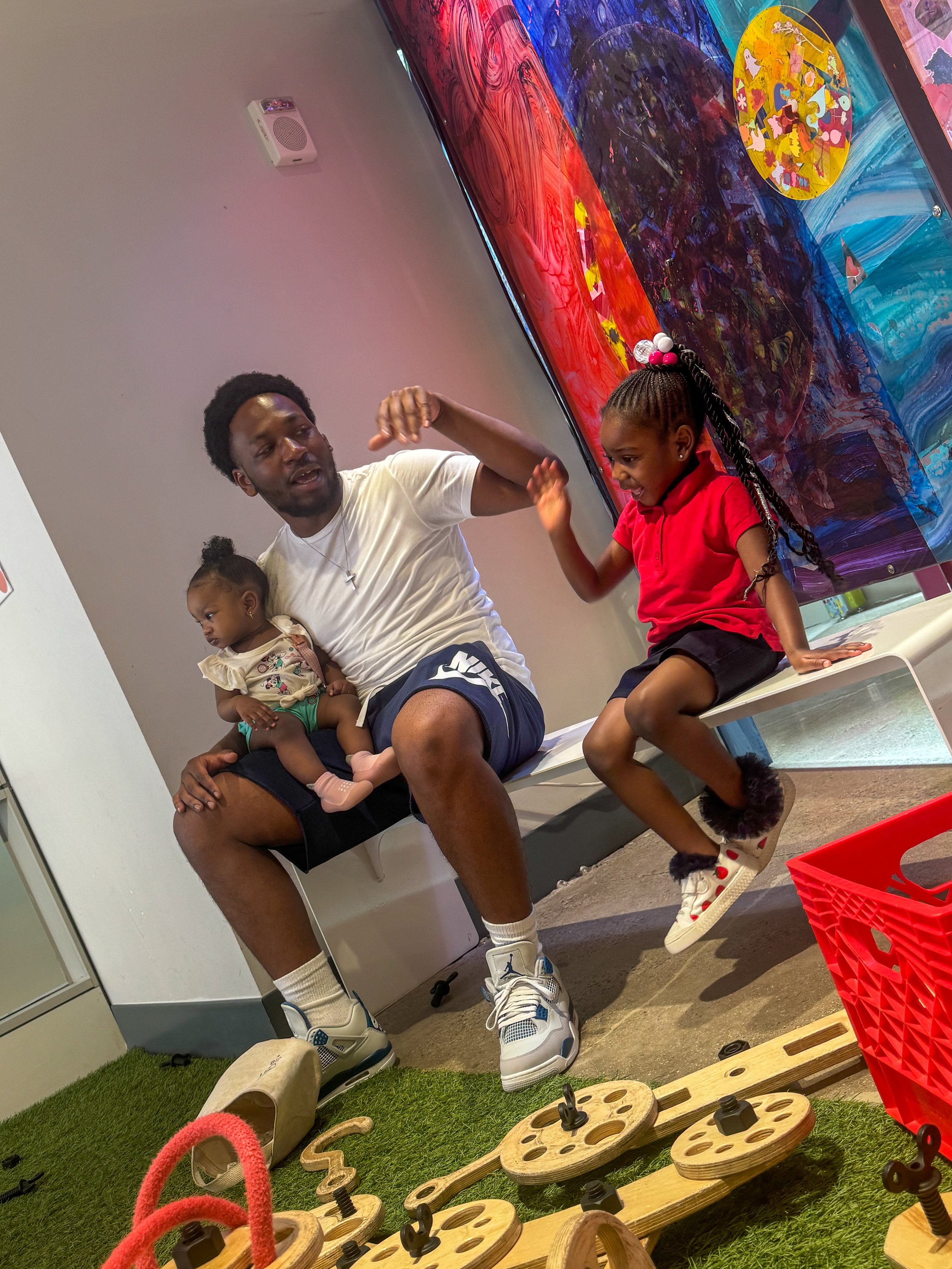A man with two young girls sitting on a bench in an indoor play area with colorful artwork on the wall behind them. One girl is wearing a red shirt and black shorts, and the other girl is sitting on the man's lap. There are wooden toy car tracks and 