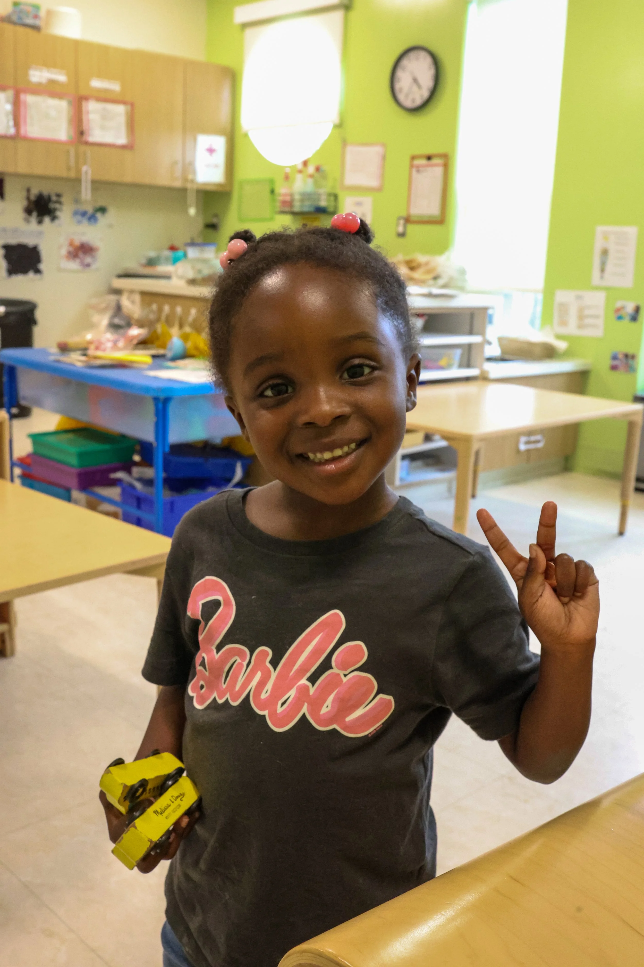 A young girl smiling and making a peace sign with her fingers in a classroom setting, holding a small yellow toy.
