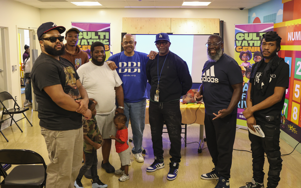 Group of seven men and three children standing together in a room with colorful walls; some are smiling, wearing casual clothing, and pose for a photo in front of a purple banner and a screen.
