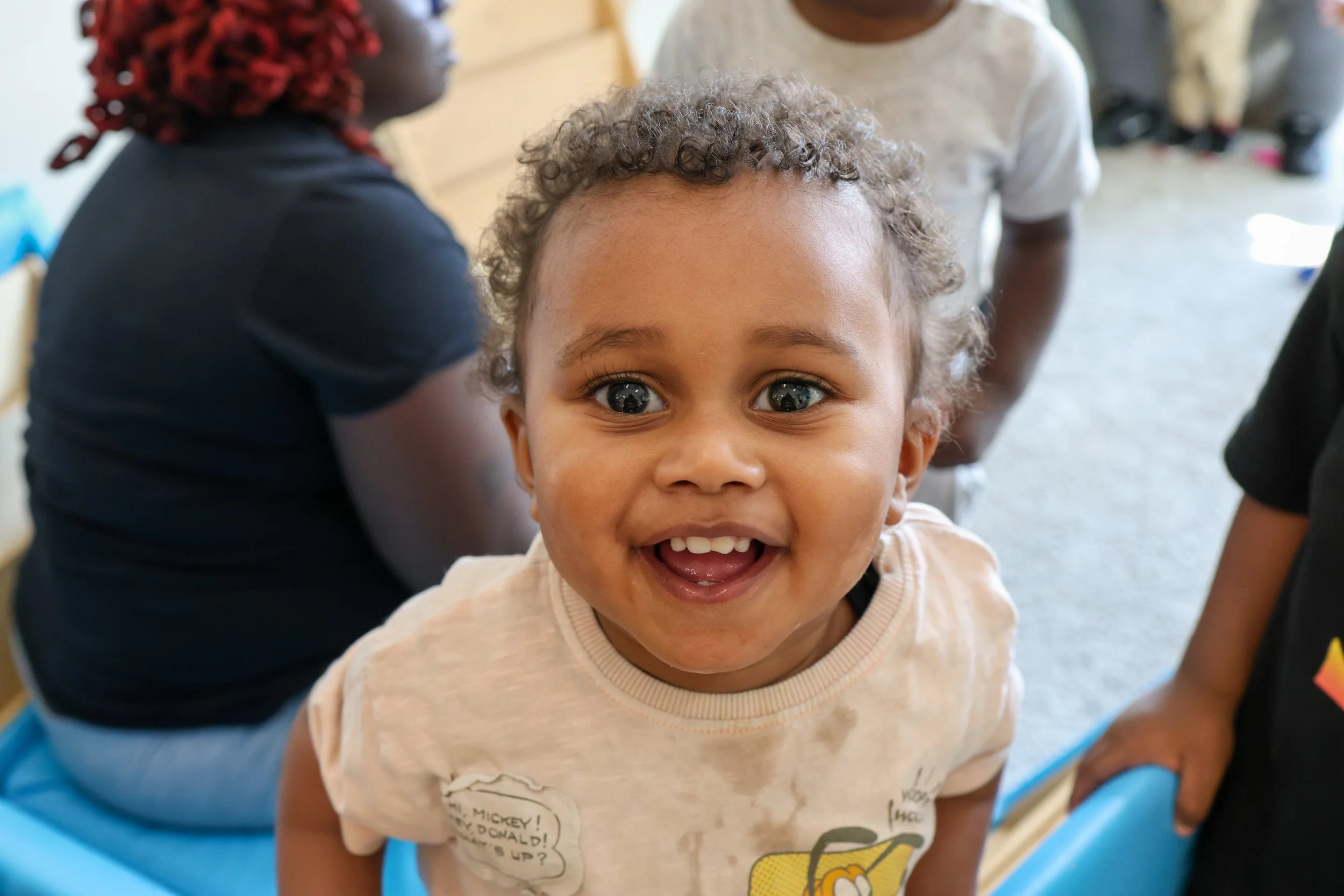 A smiling young child with curly hair and bright eyes looking at the camera, surrounded by other children in an indoor setting.