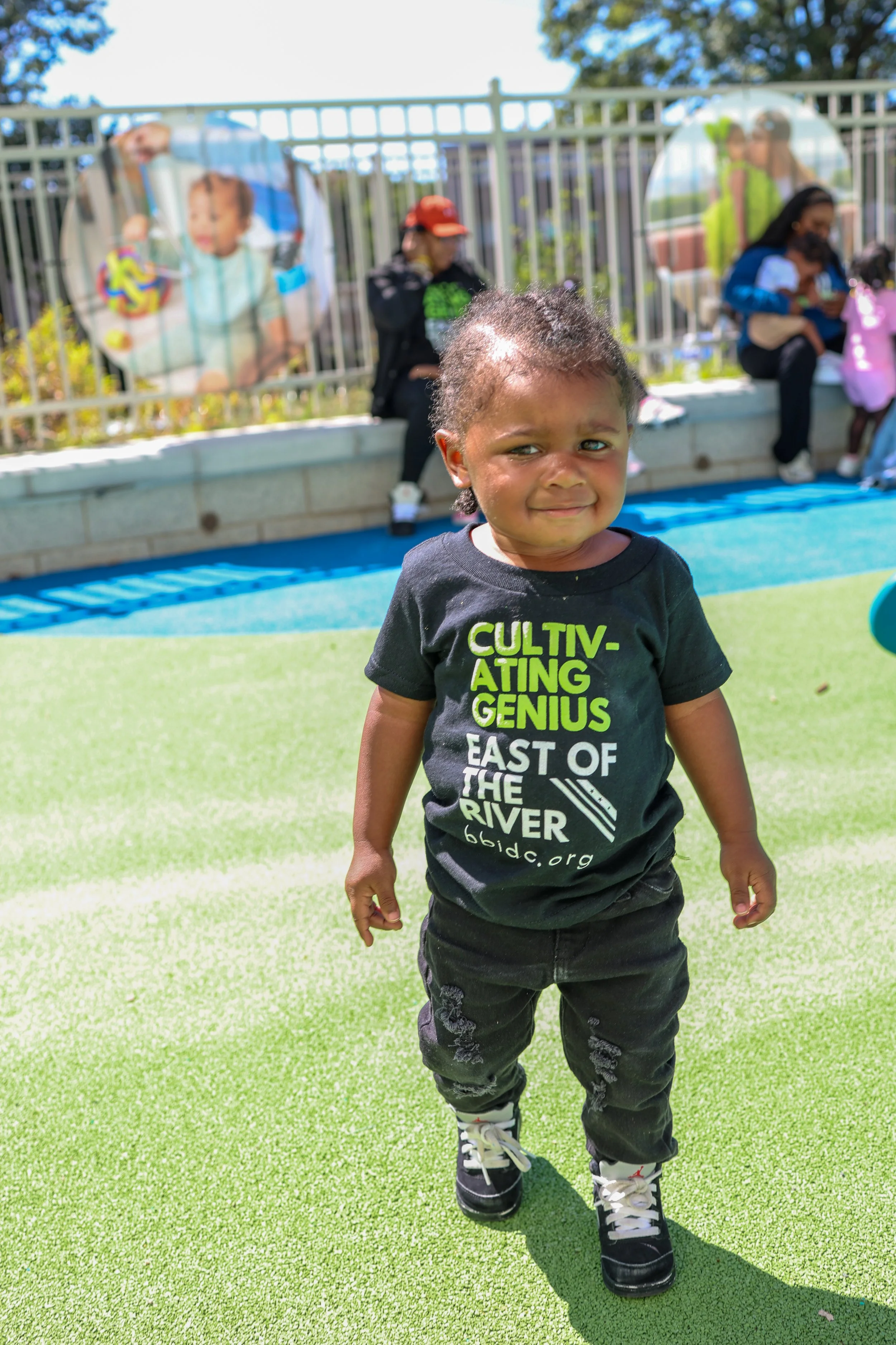 A young child standing outside on a sunny day, wearing a black T-shirt with green and white text and design, distressed black pants, and black sneakers, with people sitting and standing behind a fence in the background.