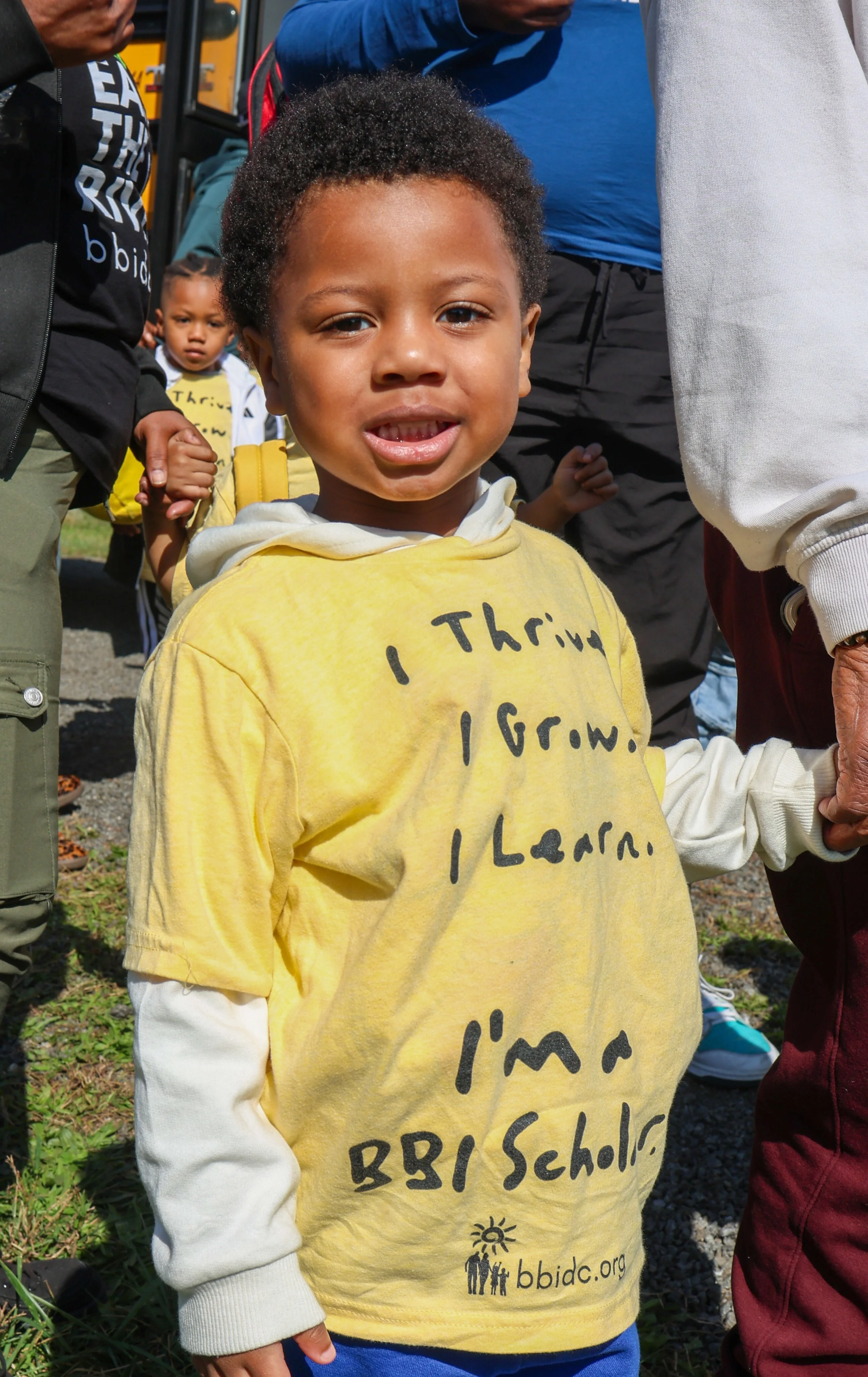 A young boy wearing a yellow shirt with handwritten words and a logo, standing outdoors among a group of people, holding hands with an adult.