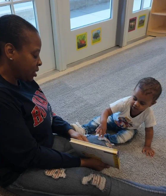 A woman and a young boy sitting on the carpeted floor indoors, engaging in reading a book together. The woman is holding the book while the boy is sitting nearby, looking at it.