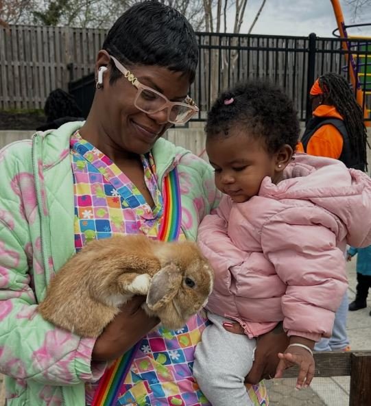 Woman holding a brown rabbit and a young girl with a pink jacket at an outdoor park or playground.