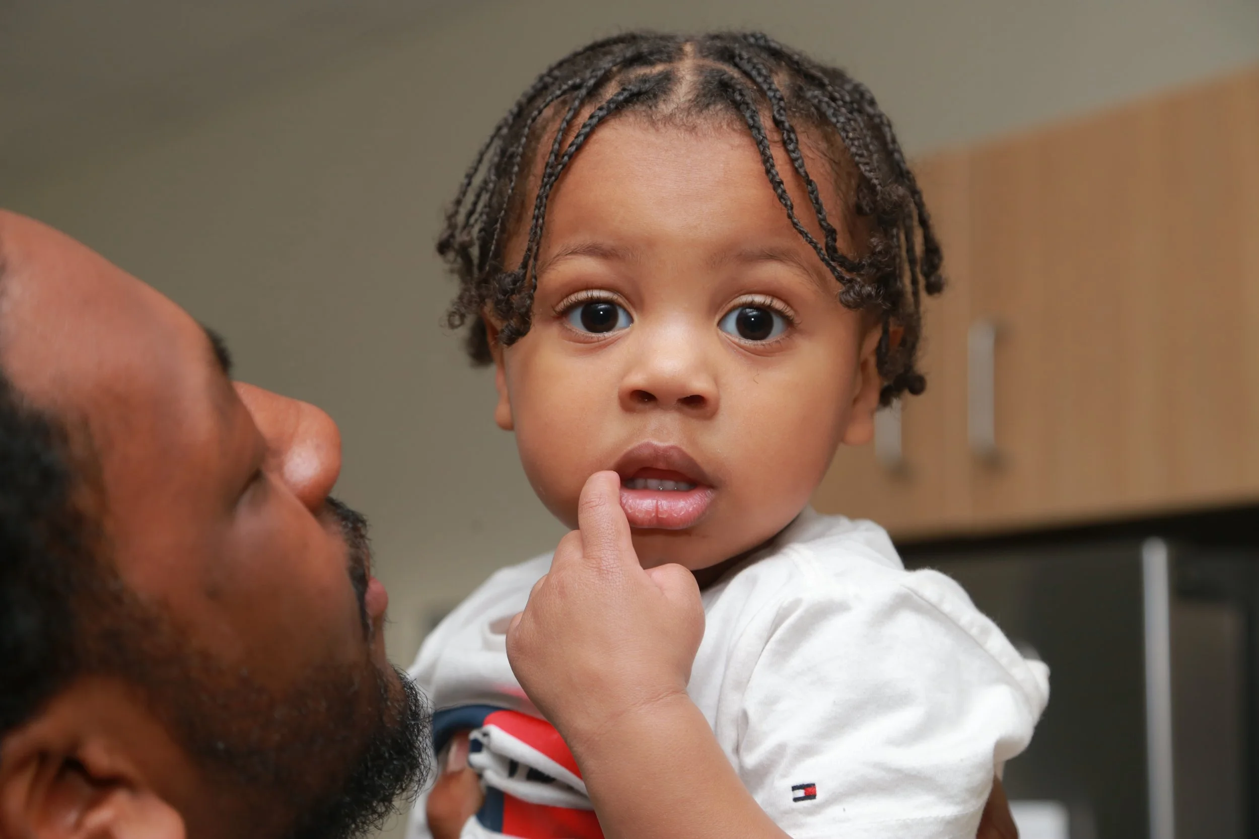 A man holding a young boy with braided hair and big eyes, touching his chin with his finger, in an indoor setting.