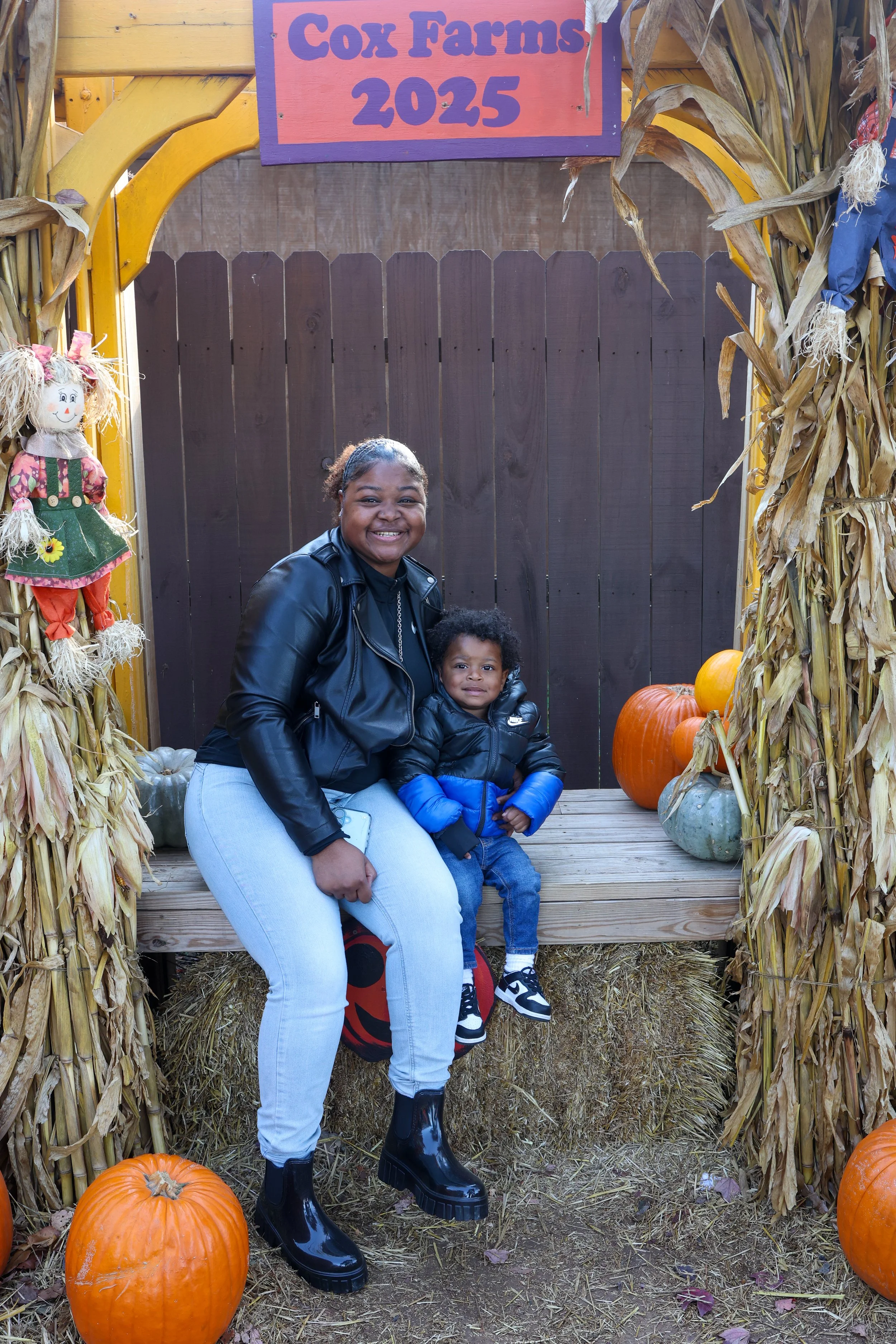 A smiling woman and young boy sitting on a wooden platform, surrounded by pumpkins and autumn decorations, under a sign that reads "Cox Farms 2025".