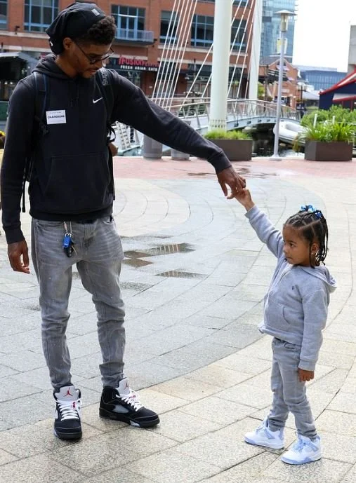 A young girl holding the hand of an older boy or man, walking outdoors in an urban area with modern buildings and a bridge in the background.