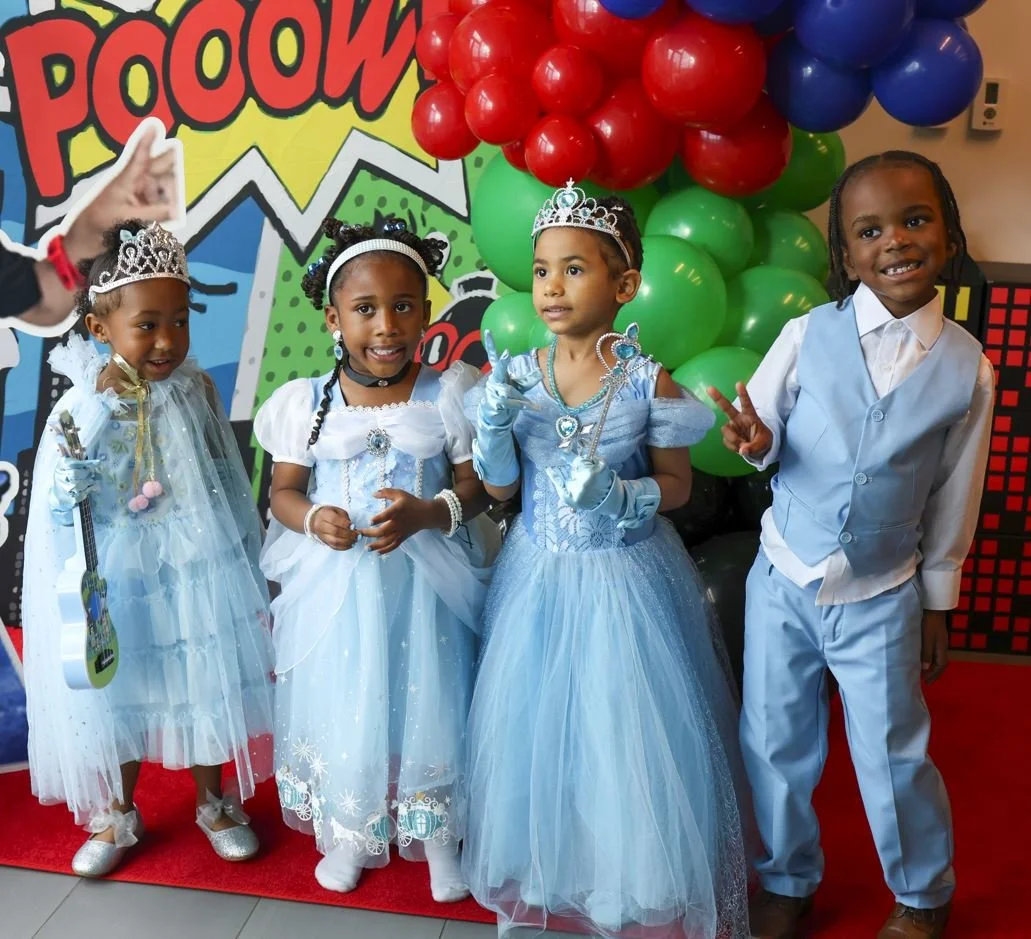 Four young children dressed as princesses and a boy at a birthday celebration, with colorful balloons and a comic book style backdrop in the background.