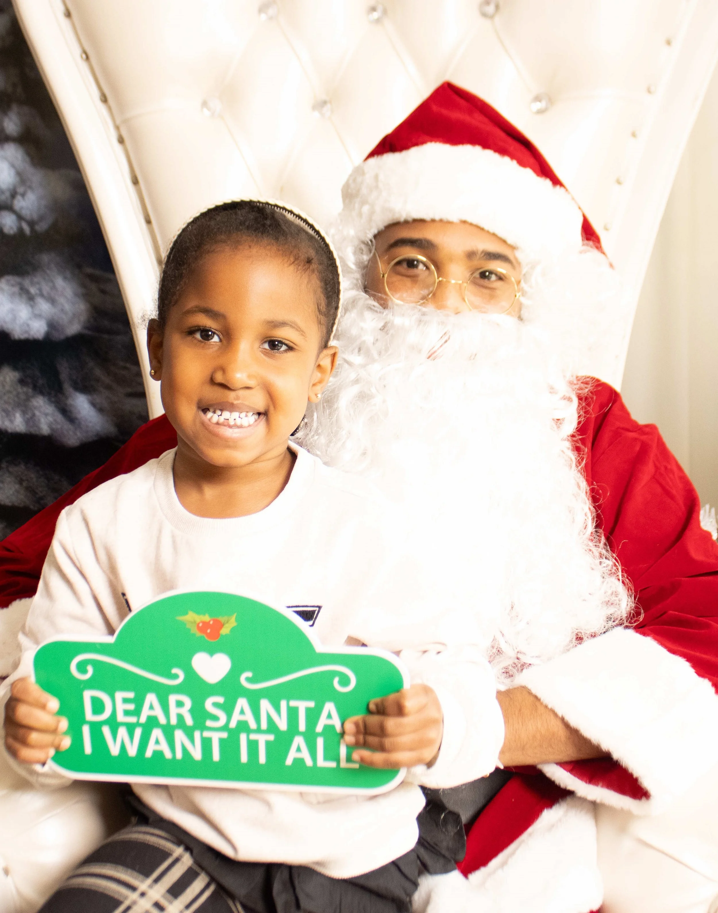 A young girl sitting on Santa Claus's lap, holding a green sign that reads 'Dear Santa I Want It All,' with Santa dressed in traditional red and white outfit with a Santa hat, a white beard, and glasses.