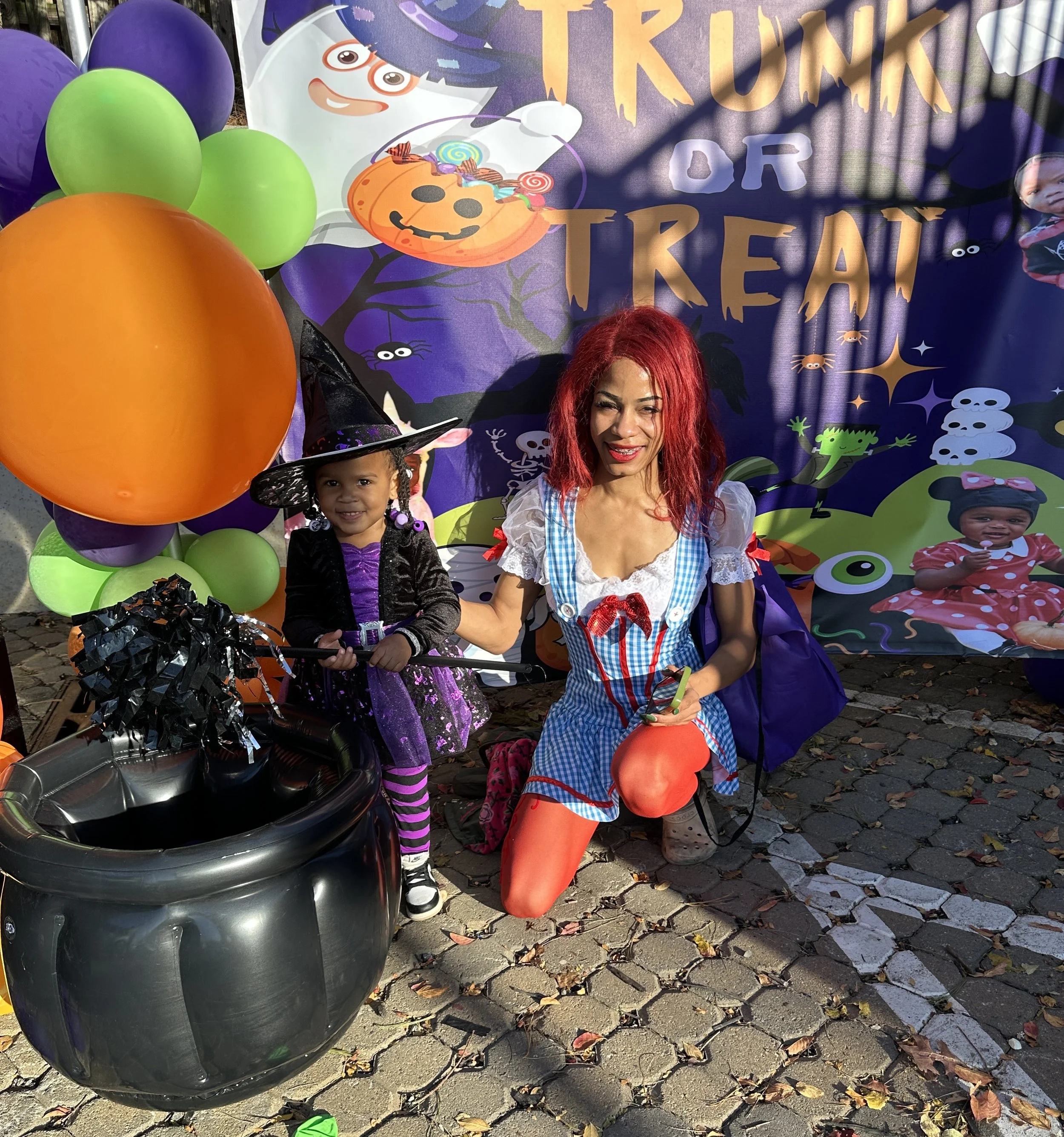 A woman and a young girl dressed in Halloween costumes posing in front of a Halloween-themed backdrop with balloons, a witch hat, a black cauldron, and decorations. The woman is dressed as Dorothy from The Wizard of Oz and the girl is dressed as a wi