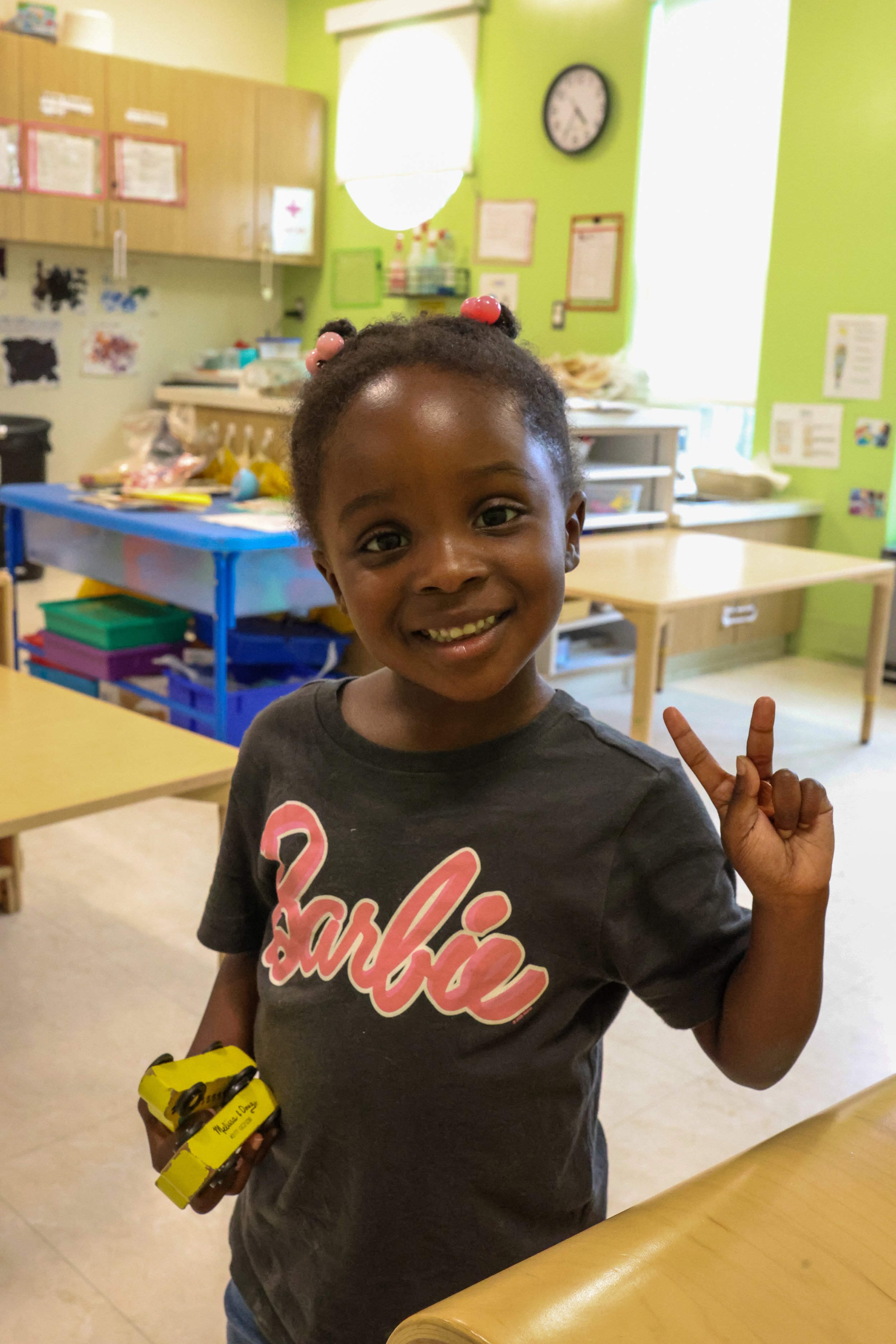 A smiling young girl in a classroom making a peace sign with her right hand while holding a yellow box in her left hand, with colorful classroom furniture and green walls in the background.