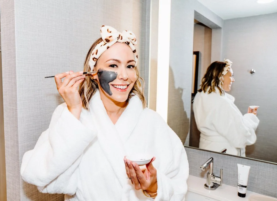 A woman in a white bathrobe and a polka dot headband applies a facial mask with a brush in a bathroom, smiling at the mirror.