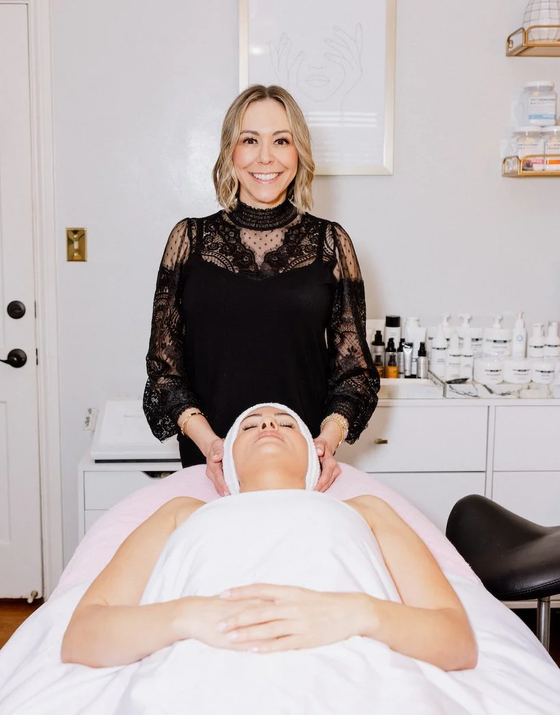A woman lying on a massage or skincare table with a towel over her chest, receiving a facial or spa treatment, while another woman stands behind her, smiling and holding the client's head. The setting appears to be a spa or skincare clinic with skincare products on shelves in the background.