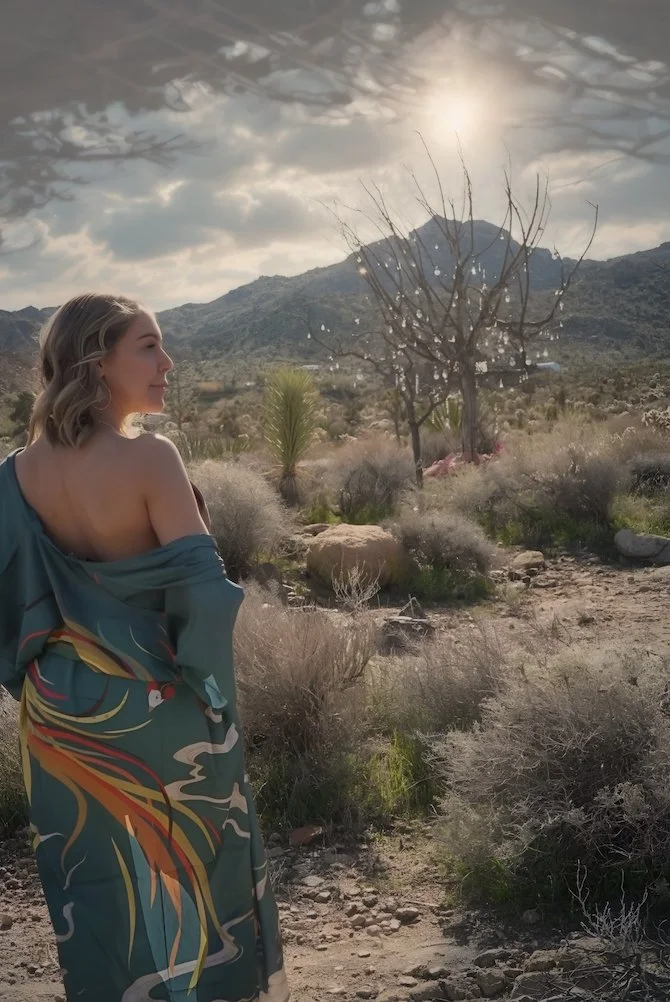 A woman in a colorful kimono stands in a desert landscape with mountains in the background and a cloudy sky overhead.