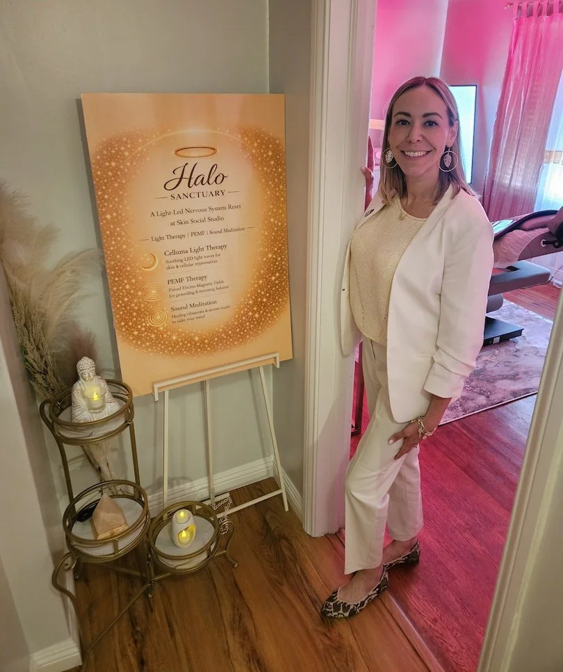 A woman in an off-white suit standing next to a sign for Halo Sanctuary, which offers light therapy, sound meditation, and PEMF therapy. She is smiling and standing on a wooden floor near a door with pink curtains in the background.