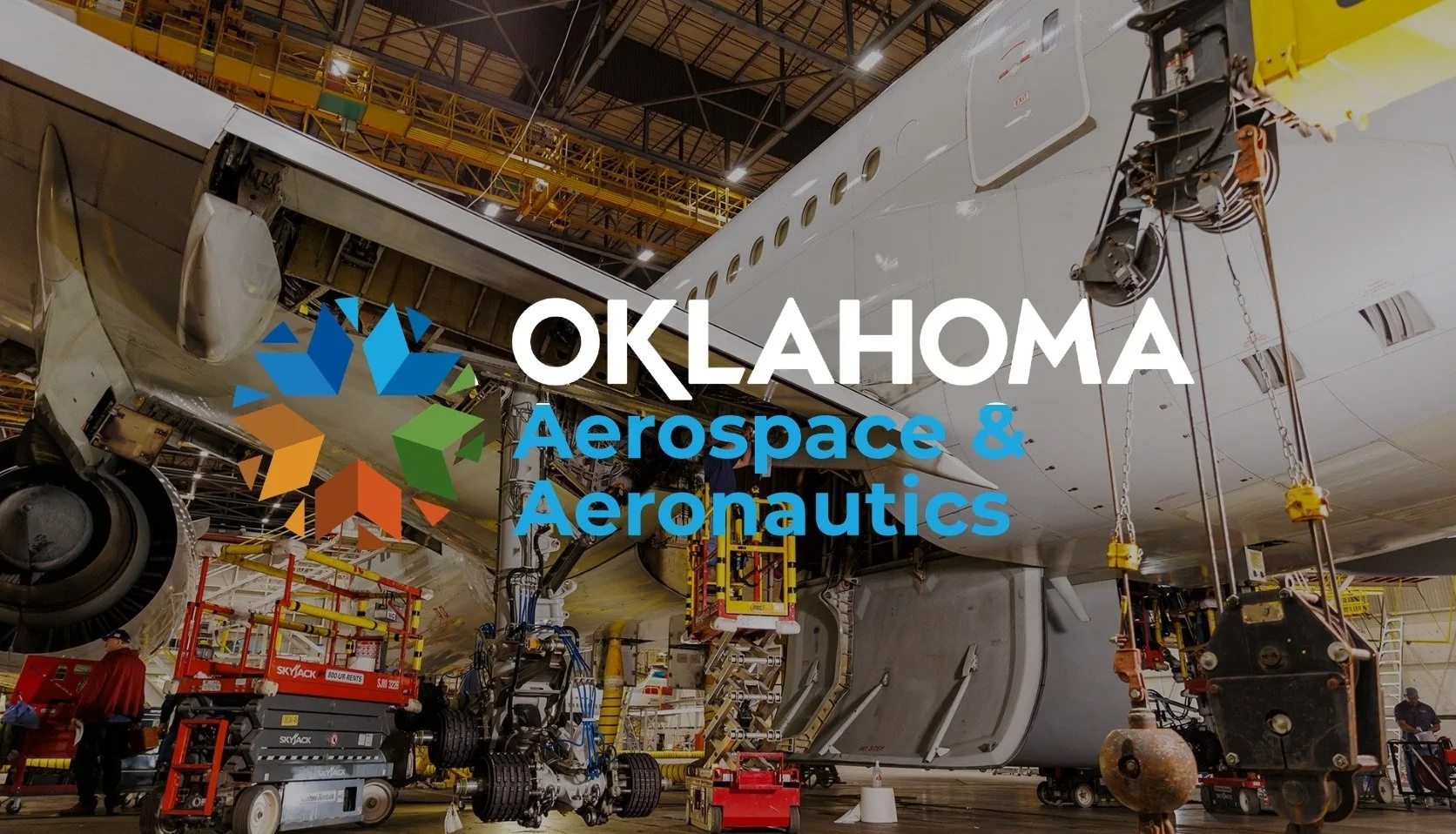 Inside an aerospace manufacturing hangar with an airplane being assembled, with equipment and workers visible.
