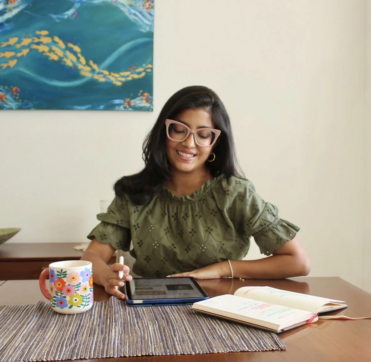 A woman with dark hair, wearing glasses and a green blouse, sitting at a wooden table with a colorful mug, an open notebook, and a tablet. A painting of dolphins swimming in the ocean is on the wall behind her.