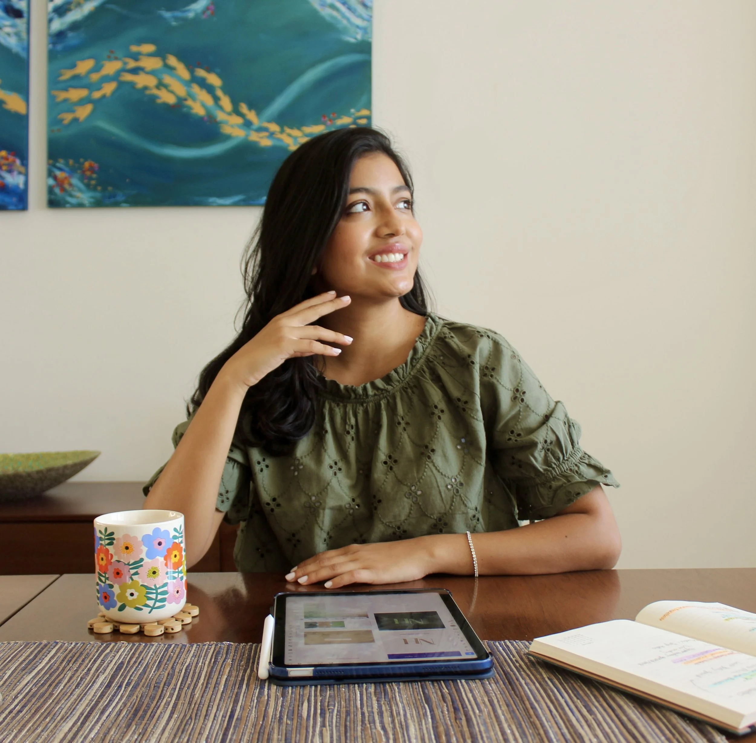 A woman sitting at a table with a tablet, an open notebook, a colorful floral mug, and a decorative bowl behind her, looking to the side and smiling.