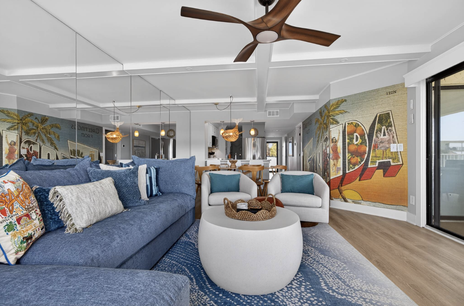 Living room with a blue sofa, white armchairs, a coffee table with a basket, wooden dining table, large wall mural with colorful Caribbean-themed art, hardwood floors, ceiling fan, and sliding glass door.