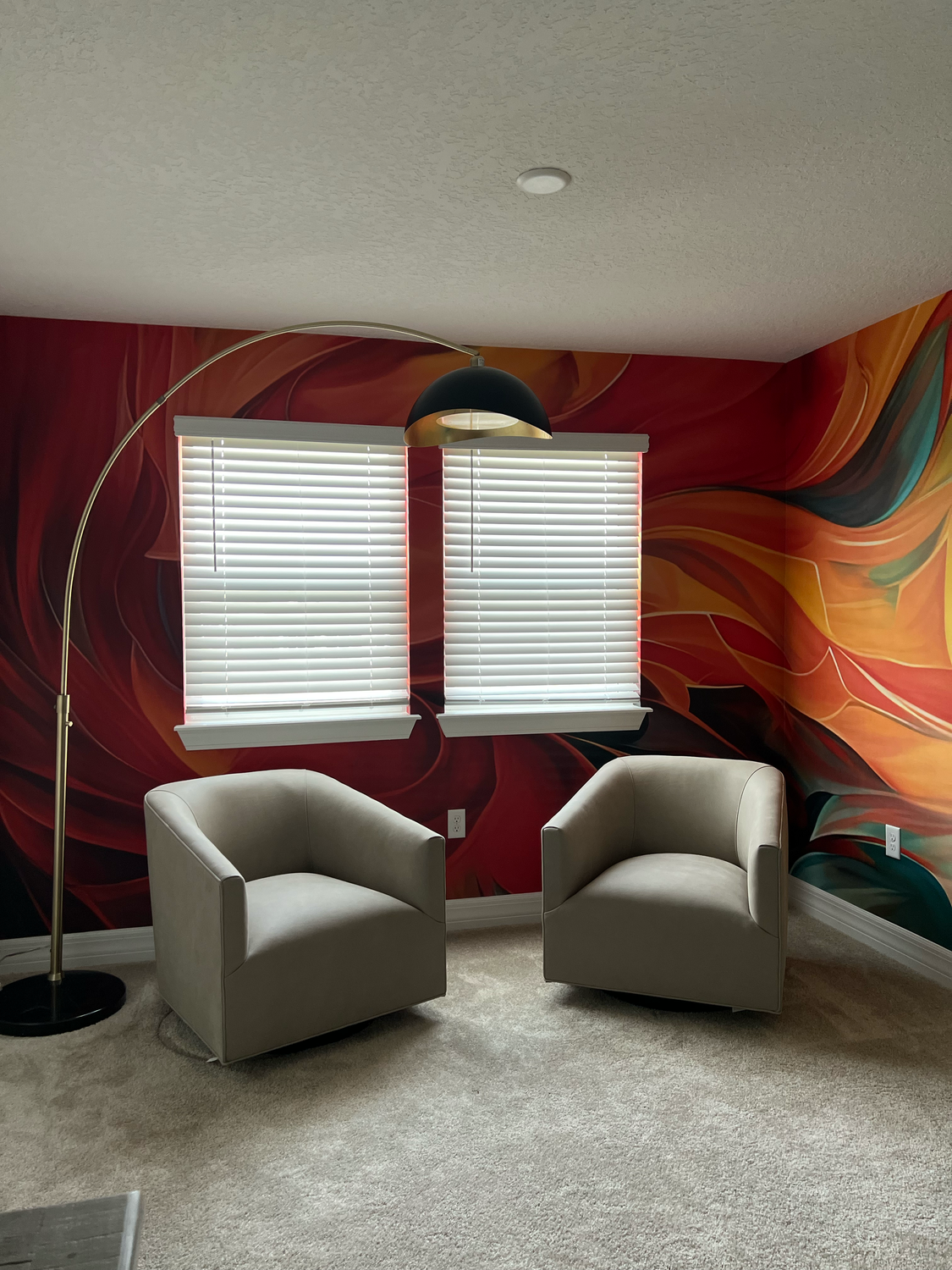 A cozy corner with two beige armchairs, large abstract floral wallpaper, a black arc floor lamp, and closed white blinds covering two windows.