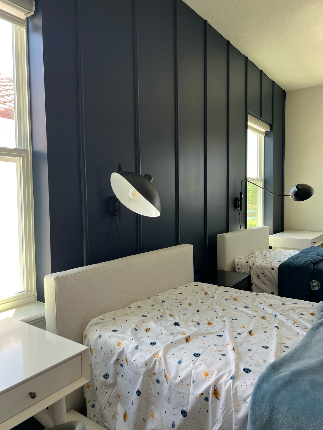 Two twin beds with beige upholstered headboards, white nightstands, and space-themed bedding in a modern bedroom with dark blue panel wall and two adjustable black wall lamps.