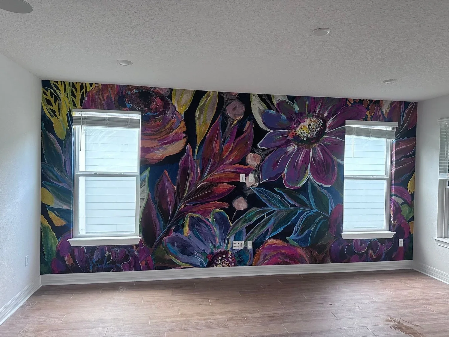 Interior room with a vibrant floral mural on the wall, two windows with white blinds, and a wooden floor.