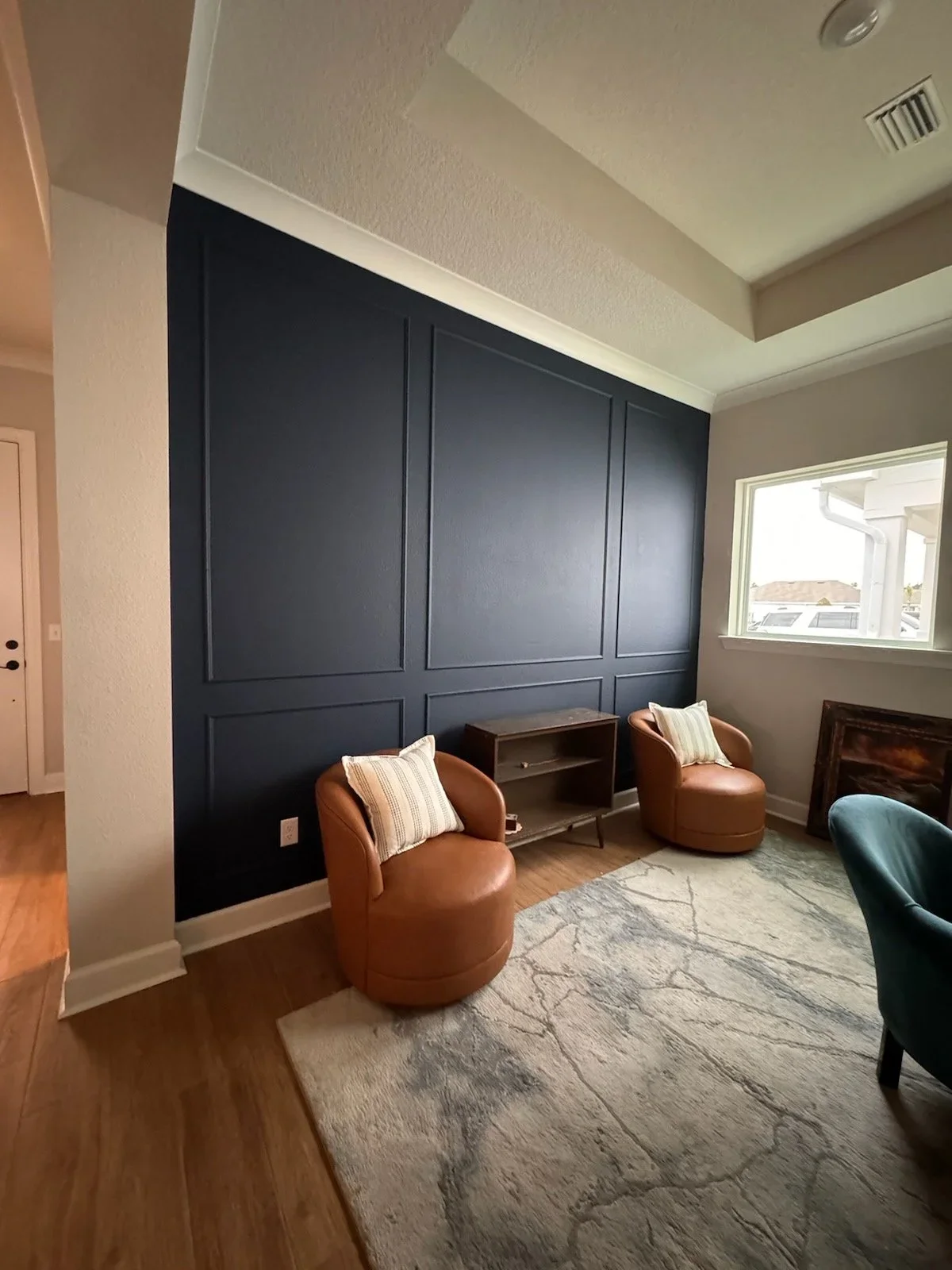 Living room with dark blue accent wall, two tan leather armchairs with striped pillows, a small wooden shelf, a large window, a painting leaning against the wall, and a beige rug on wood flooring.