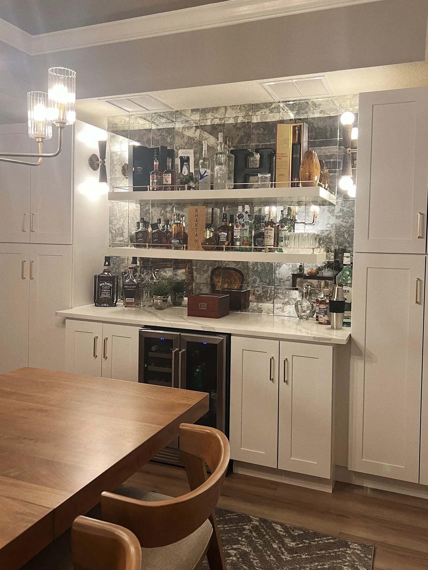 A kitchen bar area with shelves holding liquor bottles, glasses, and decorative items. White cabinets and marble backsplash, a wooden countertop, and a dining table with chairs in the foreground.