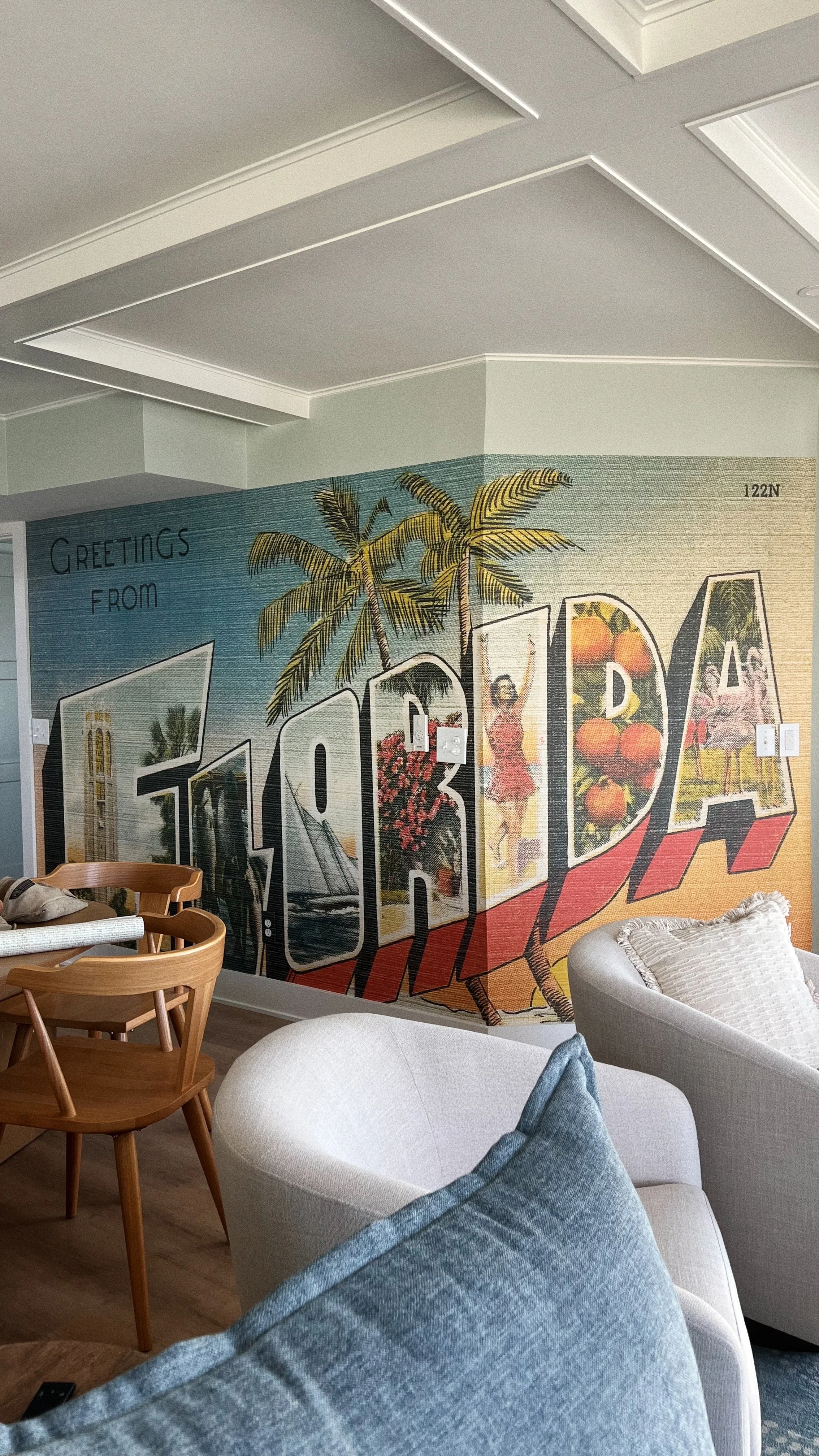 Decorative wall mural with palm trees, flowers, and the word 'Tropical' in large letters, with a partial view of chairs and a sofa in a living room.