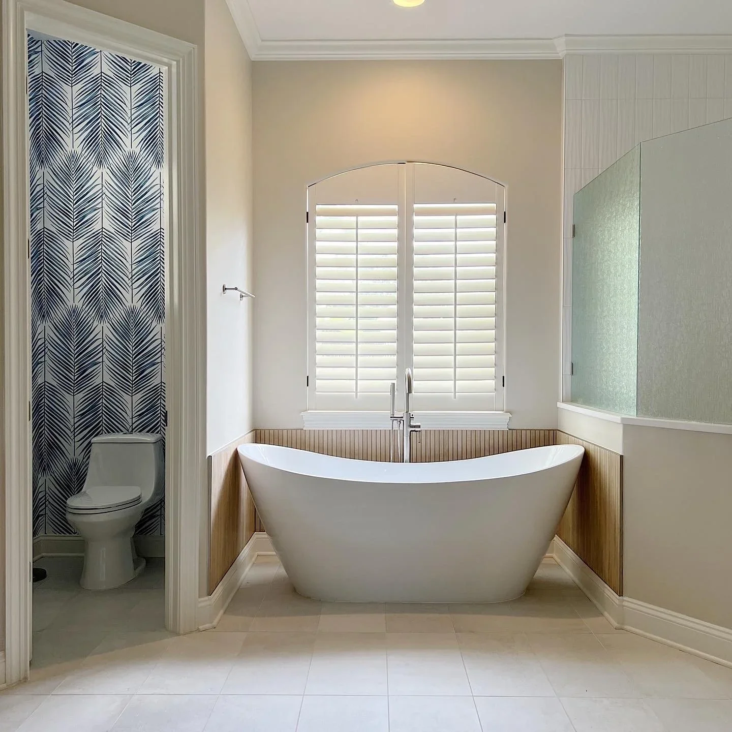 Modern bathroom featuring a freestanding bathtub positioned in front of a large window with white plantation shutters, a separate toilet area with blue and white patterned wallpaper, and a frosted glass shower enclosure to the right.