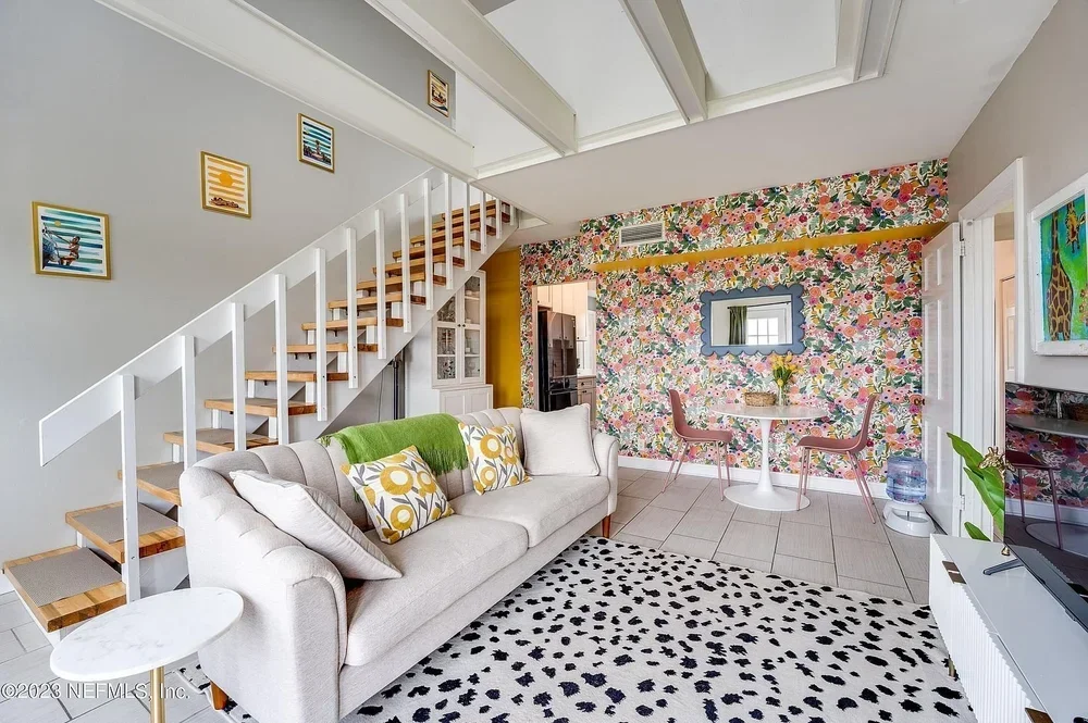 Living room with white sofa, green and patterned cushions, patterned rug, floral wallpaper accent wall, small round dining table with two pink chairs, wall art, staircase, flat-screen TV, and large windows.