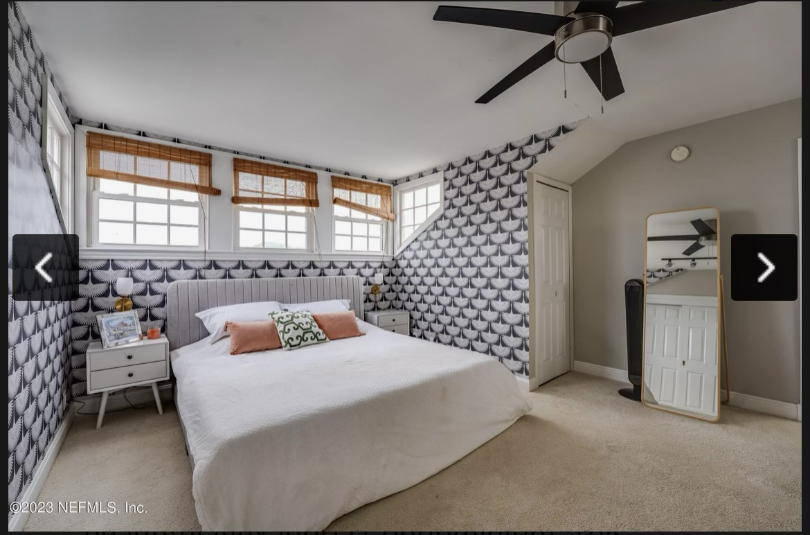 A bedroom with a bed, nightstand, window blinds, ceiling fan, mirror, and patterned wallpaper.