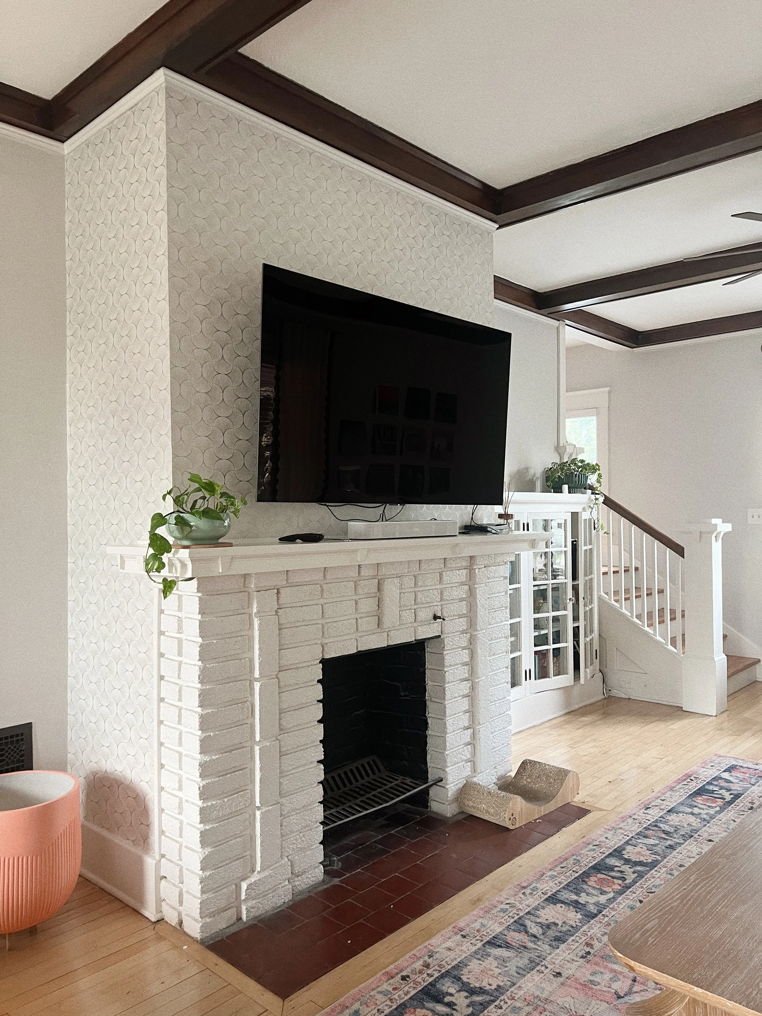 Living room with a white painted brick fireplace, a flat-screen TV mounted above the fireplace, and a white mantel with some plants and decorative objects. The room has wooden floors, patterned area rug, staircase with white railing, and partial view
