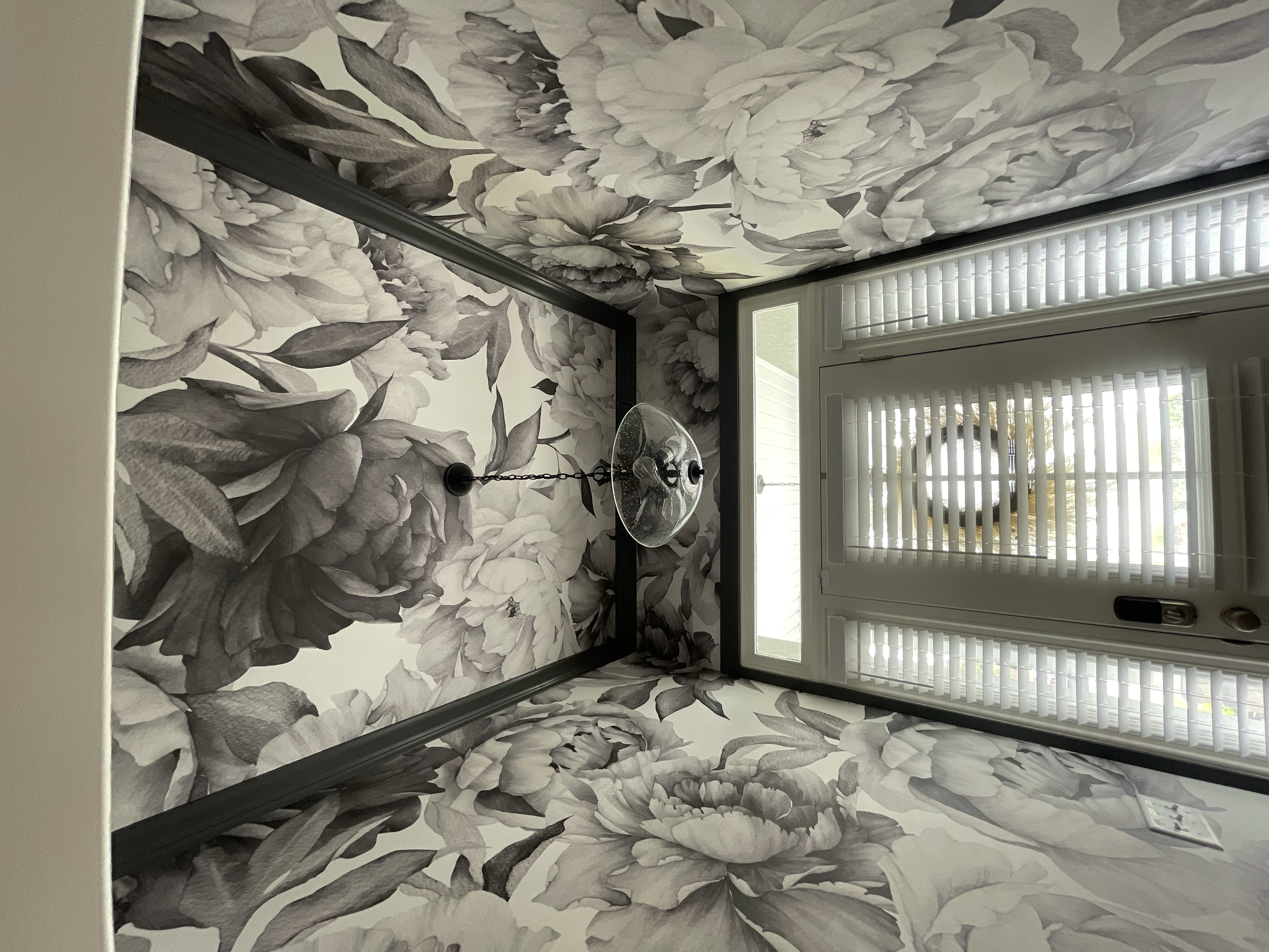 A room with black and white floral wallpaper, a door with vertical blinds, and a chandelier with light bulbs hanging from a chain.