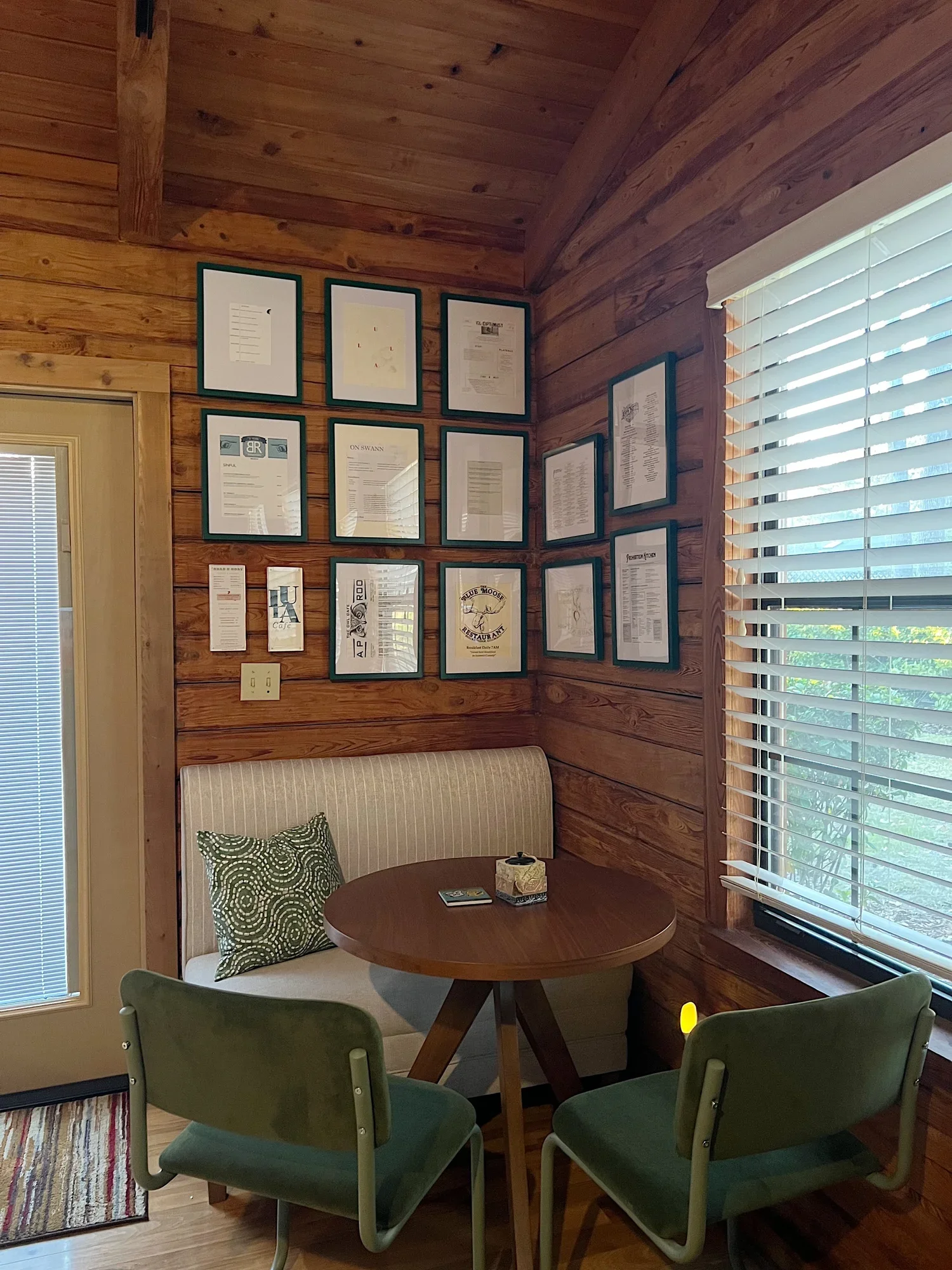 A small round wooden table surrounded by four green chairs in a cozy wooden-paneled room. A beige couch with a patterned pillow is against the wall. The wall features a collection of framed documents and artwork. To the right, there's a window with w