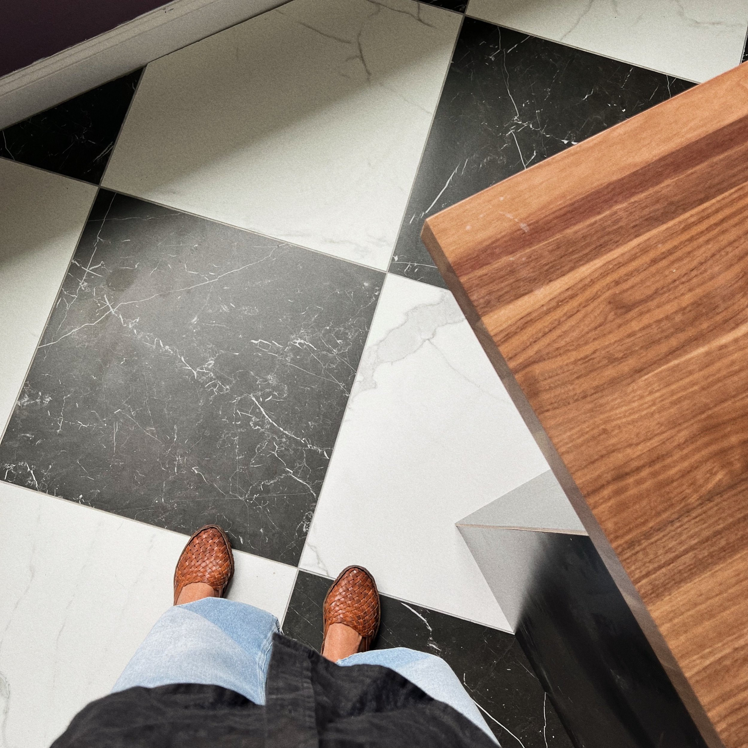 The photo shows the view from above of a person wearing woven brown shoes, blue jeans, and a black jacket, standing on a tiled floor with black, white, and grey tiles, next to a wooden piece of furniture.