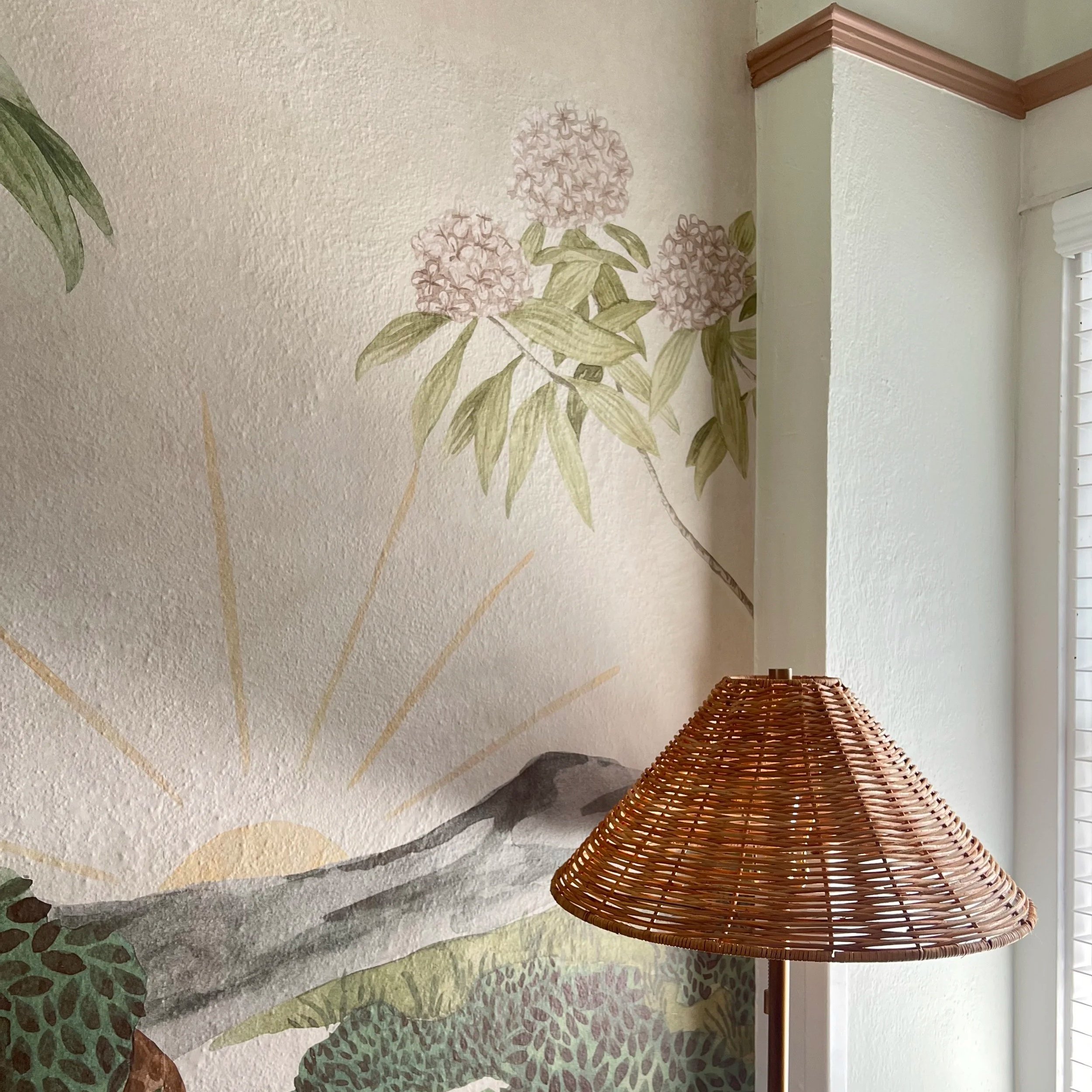 Corner of a room with textured light beige walls, a decorative floral mural, a wooden trim at ceiling, a wicker lamp in foreground, and a window with white blinds on the right.