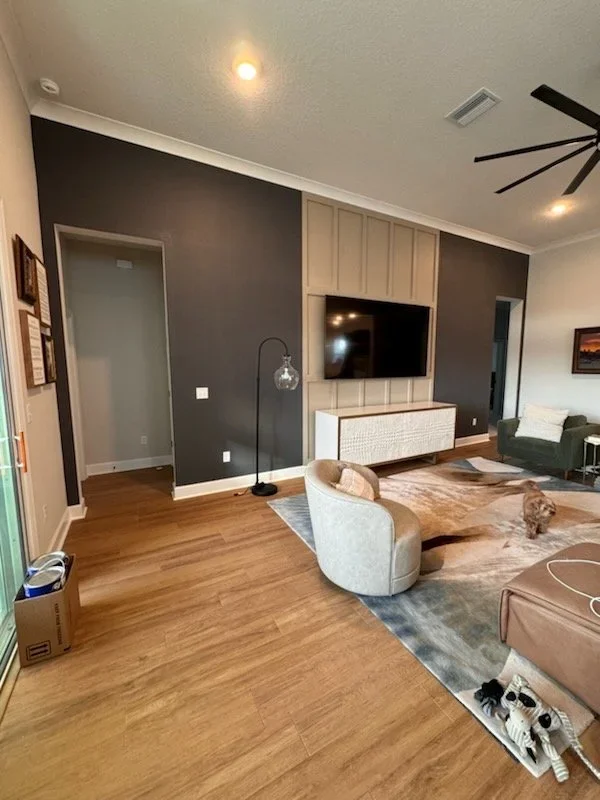 Modern living room with a dark accent wall, mounted flat-screen TV, light-colored cabinetry, gray armchair, green sofa, area rug, dog, and ceiling fan.