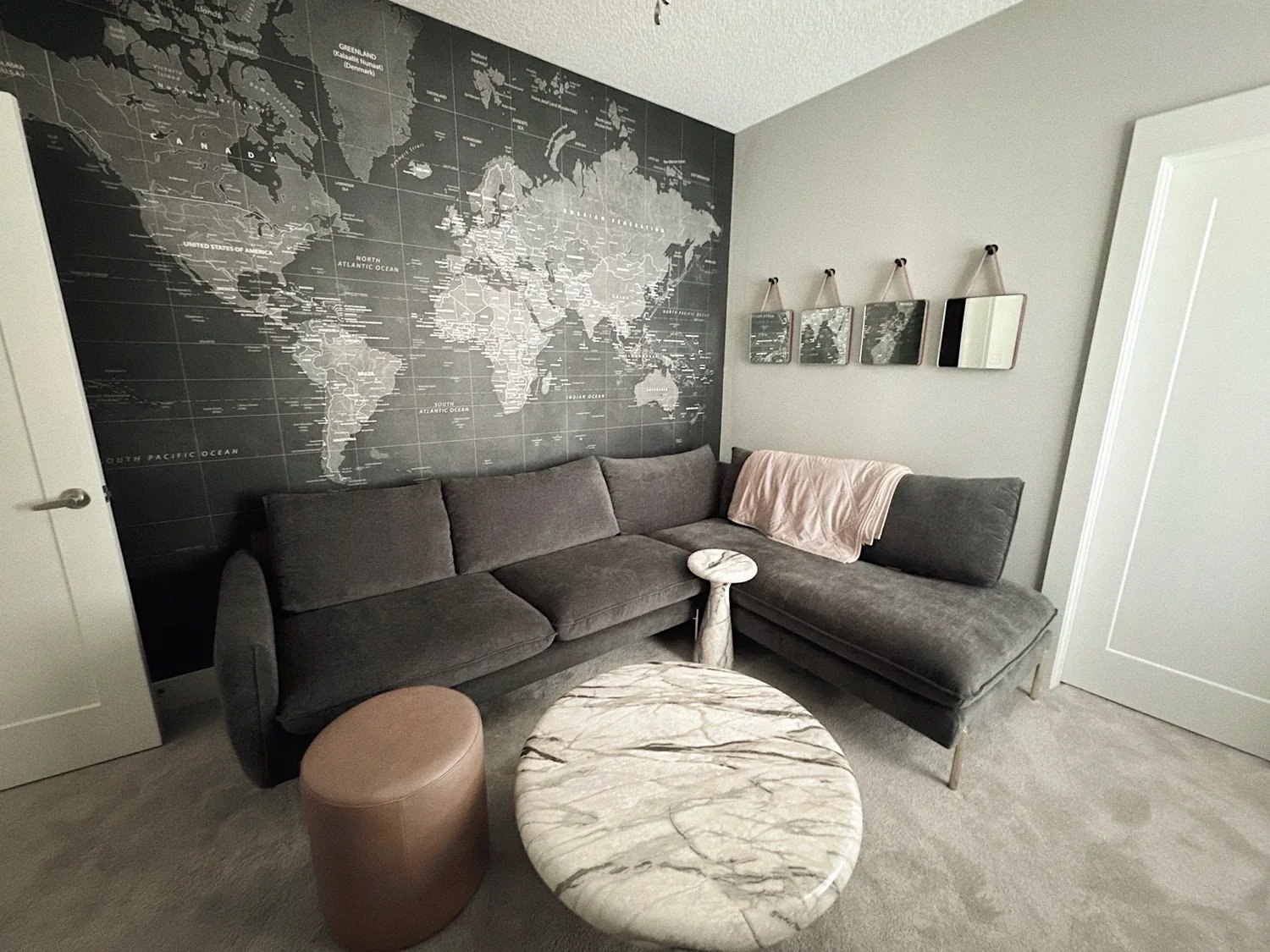 Living room with a large gray sectional sofa against a black world map wall. A round marble coffee table and a small brown ottoman are in front of the sofa. To the right, a white door and a wall with four small hanging mirrors. A pink blanket drapes 