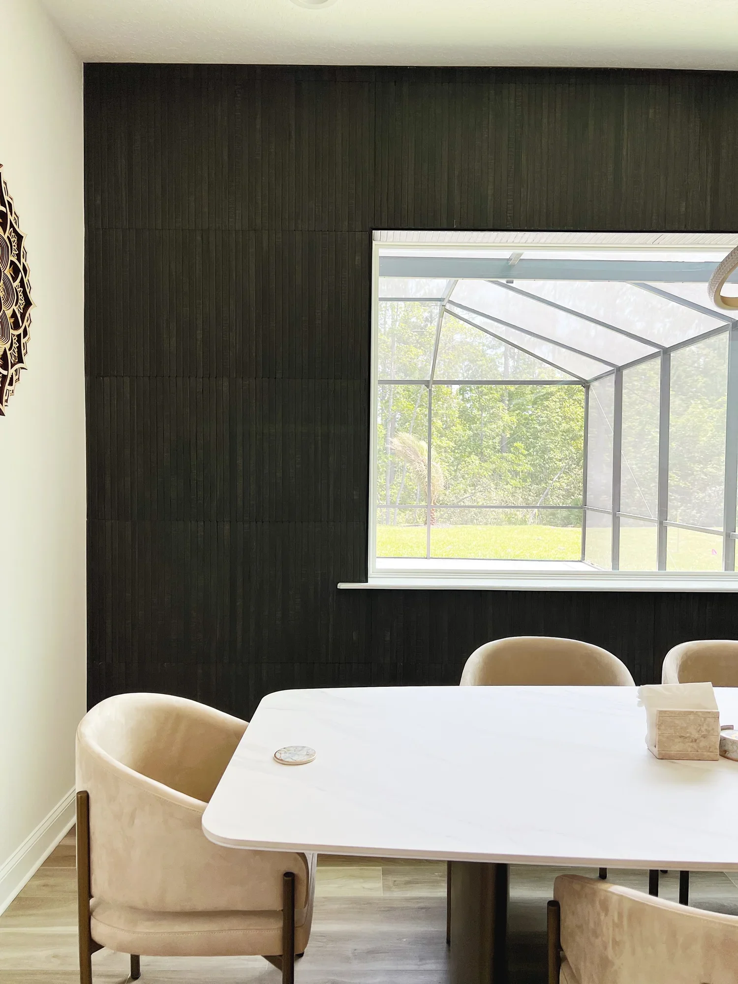 A dining area with a white table, beige chairs, and a black wood-paneled wall. A window reveals a screened-in porch and green outdoor scenery.