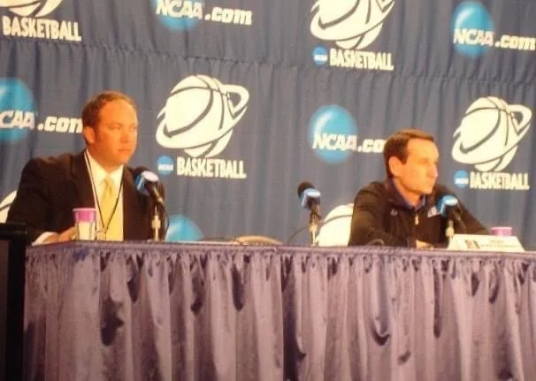 J.P. Shadrick and Duke Head Coach Mike Krzyzewski sit at a table during a press conference with NCAA and basketball logos on the backdrop.