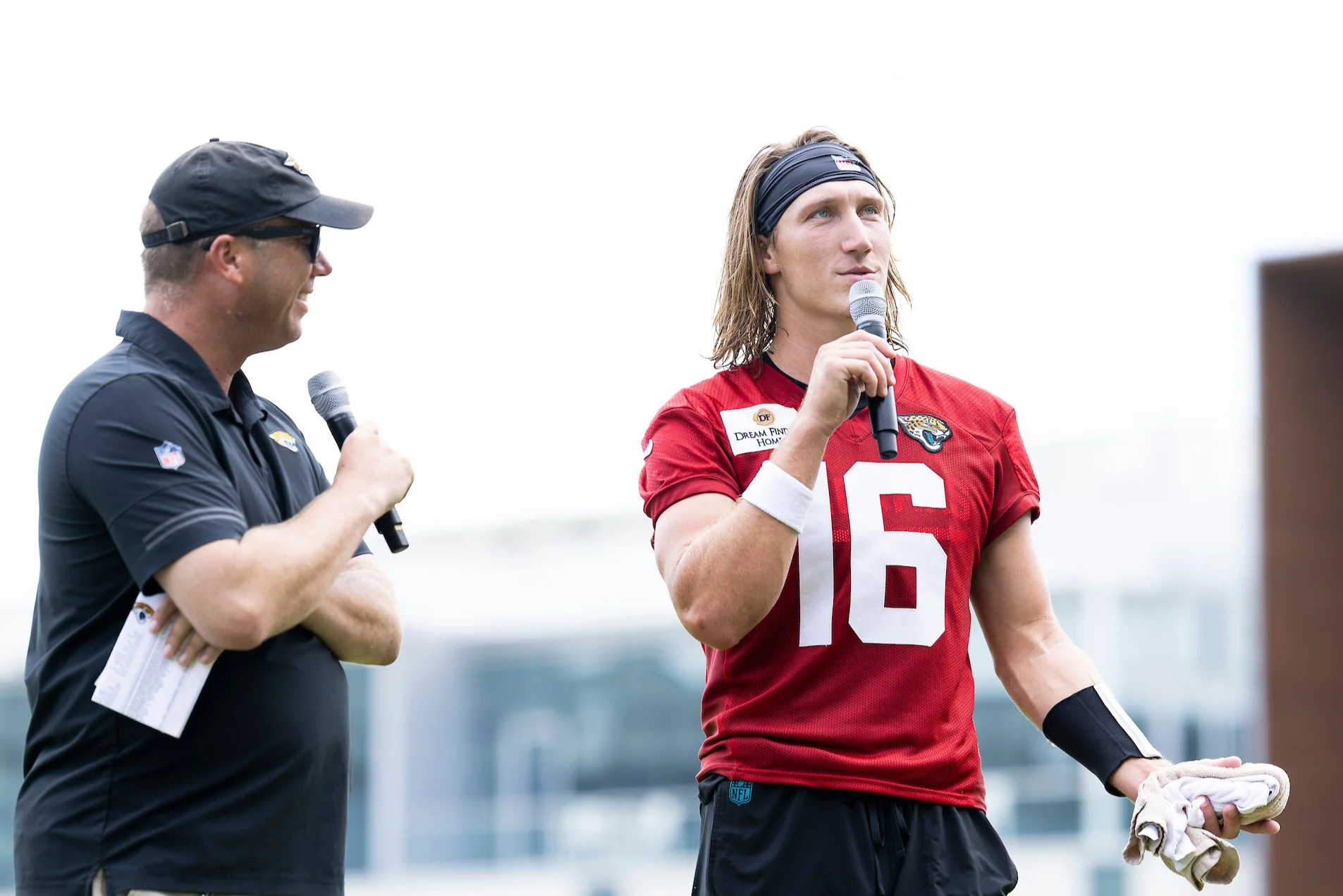 A football player in a red jersey with the number 16 talking into a microphone during a game or event, while another person, possibly a coach or team staff, in a black polo and cap listens with a microphone in hand.