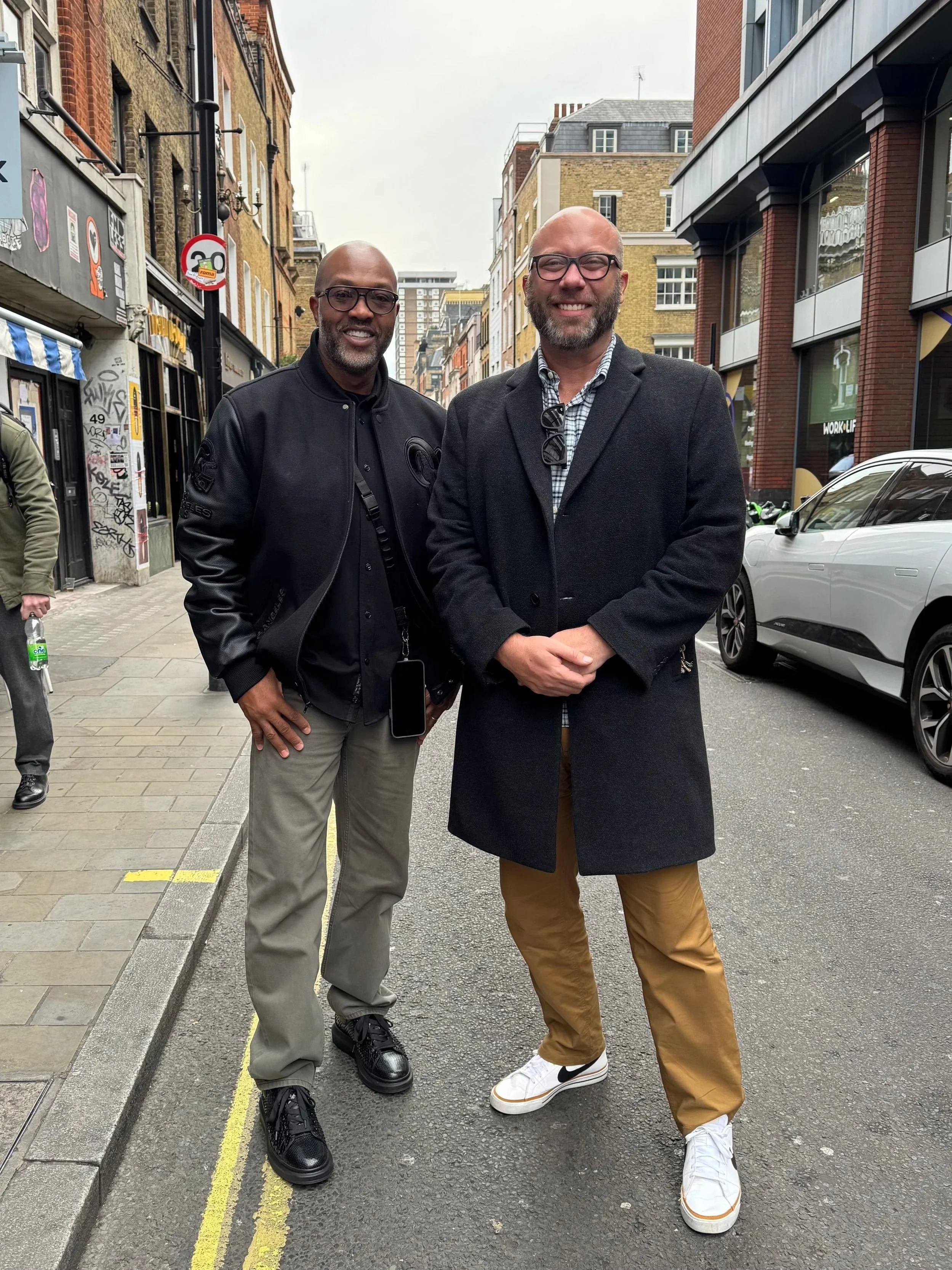Two men standing on a city street, smiling at the camera. One wears a black jacket, khaki pants, and black shoes; the other wears a long dark coat, tan pants, and white sneakers. Buildings and cars are visible in the background.