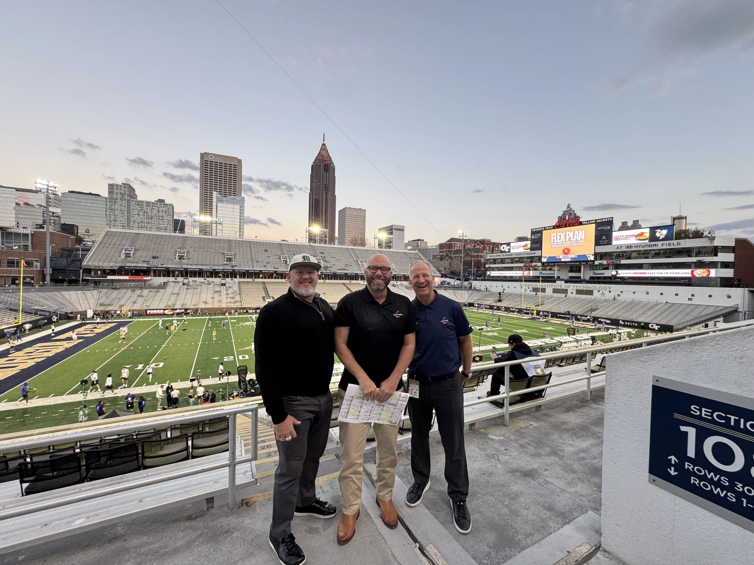 Three men standing in front of a football field at a stadium with a city skyline in the background, during sunset.