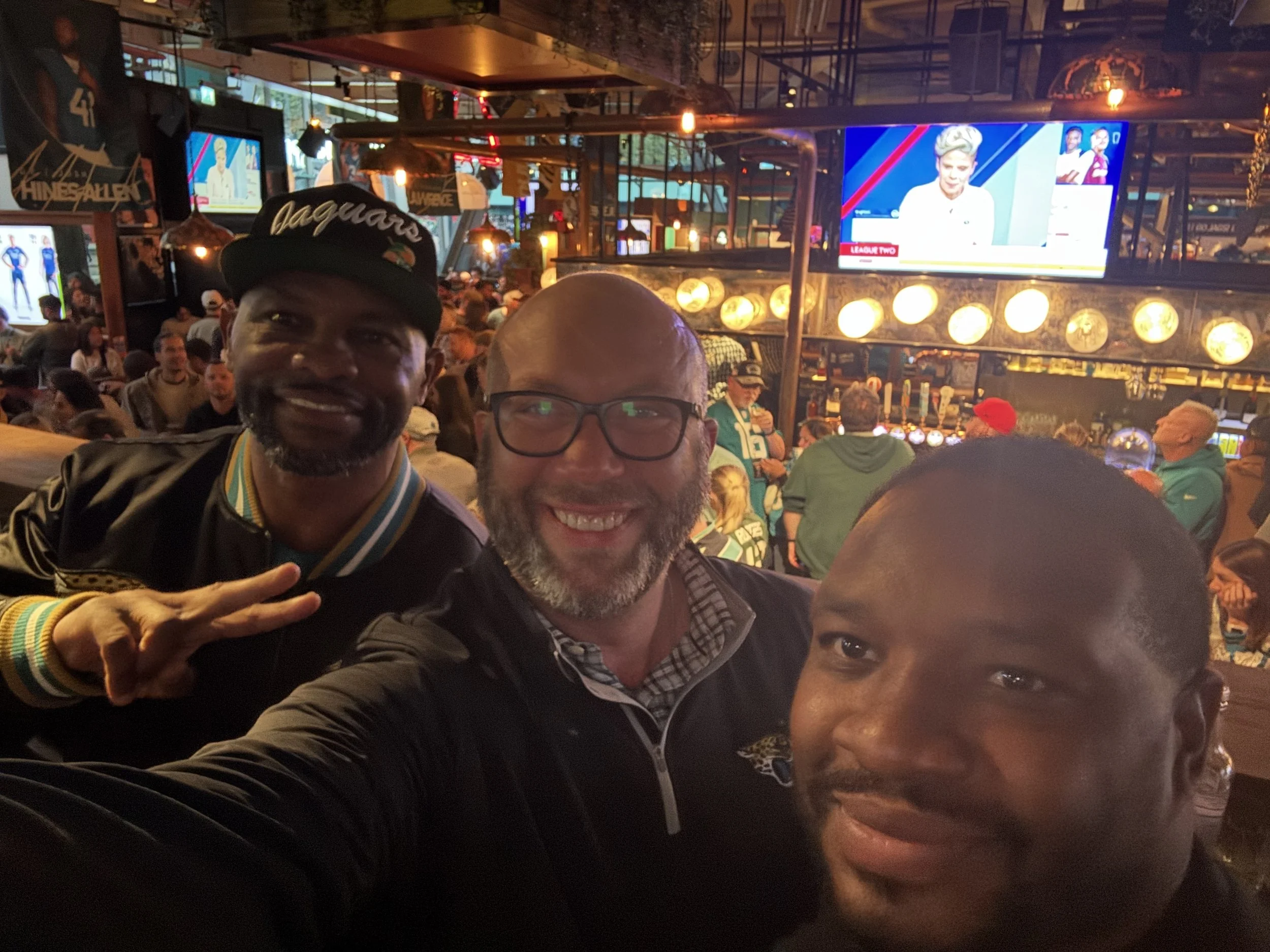 Three men taking a selfie at a crowded sports bar, with TVs showing sporting events and a bar in the background.