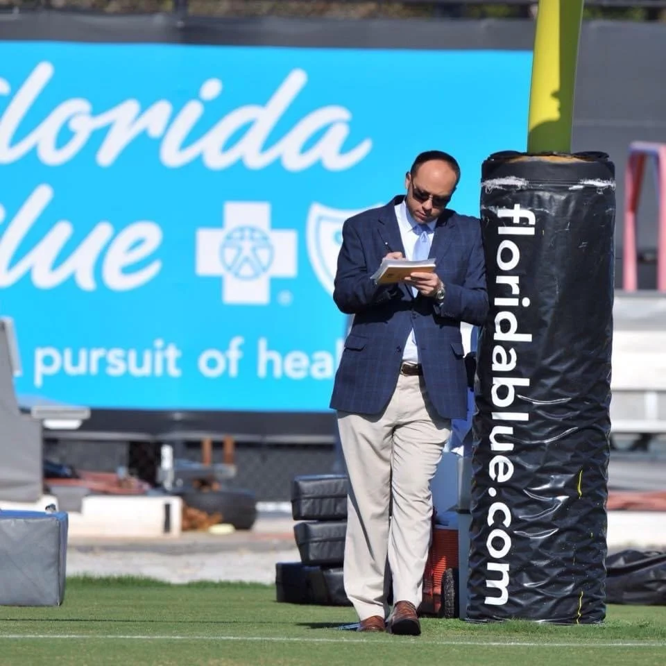 A man in a navy plaid blazer and beige trousers writing notes on a notepad outdoors next to a large safety cushion with the FloridaBlue.com logo, on a football field with a large FloridaBlue advertisement in the background.