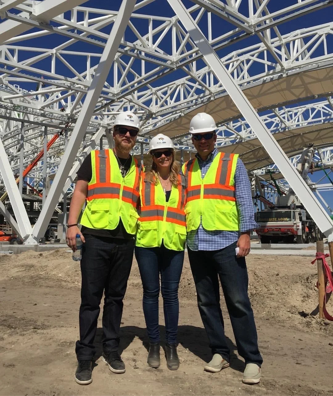 Three construction workers in safety vests and hard hats standing on dirt ground in front of a large steel structure at a construction site, under a clear blue sky.
